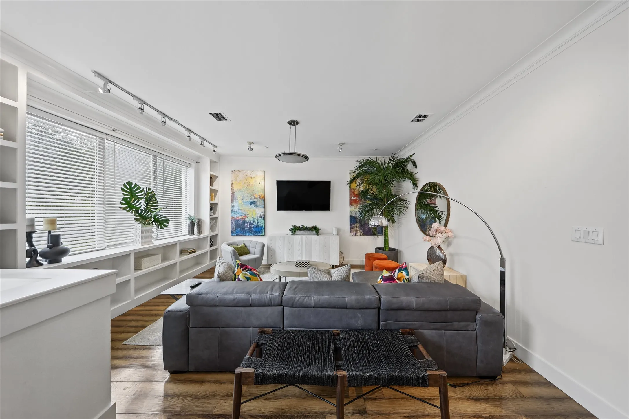 Bright, open living room showcasing hardwood flooring, a gray upholstered sectional sofa, and a wall-mounted flat-screen television above a white built-in fireplace surround