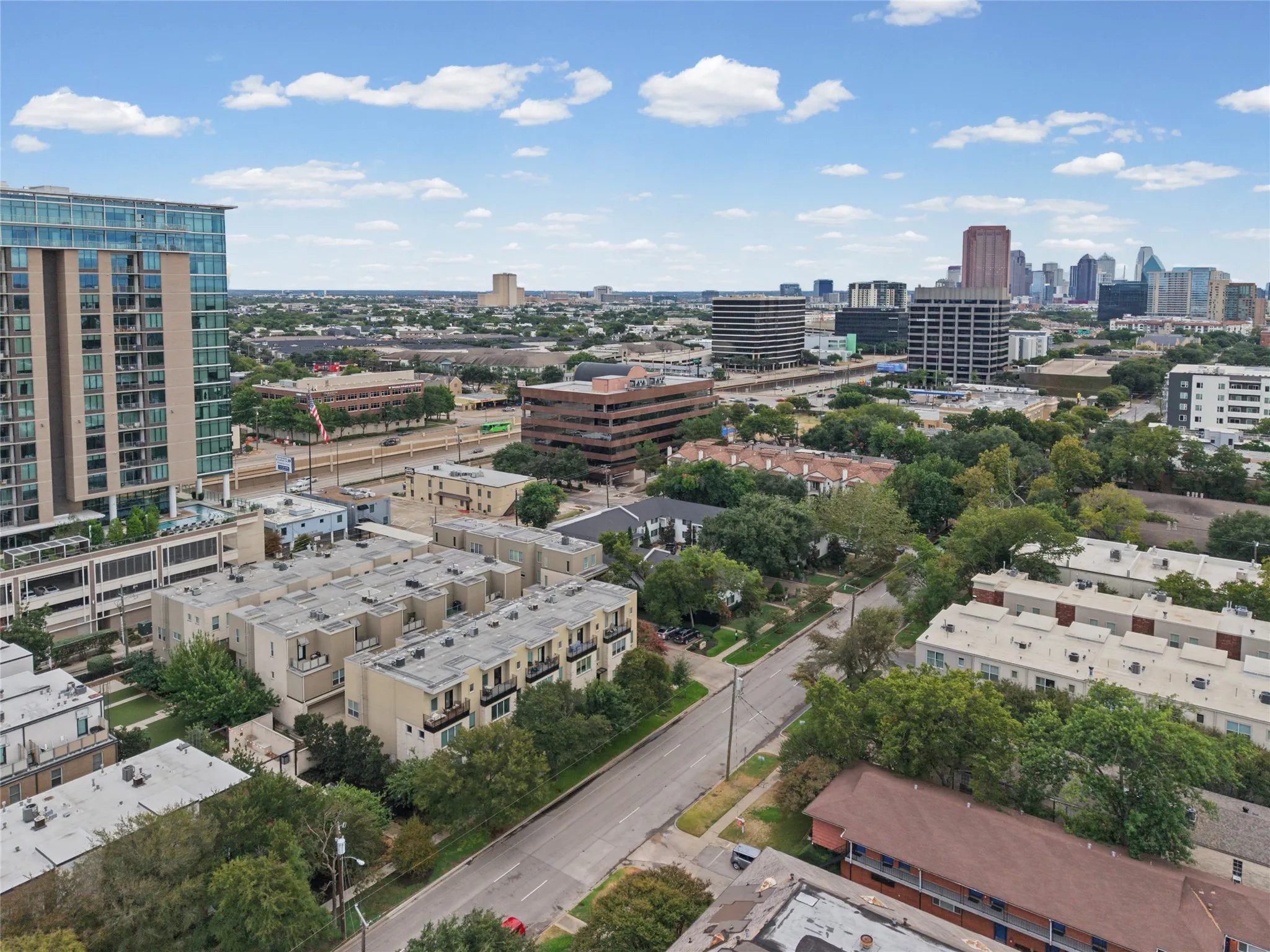 Urban landscape featuring a high-rise building with glass facade, multiple multi-story residential structures, a prominent road network, and extensive tree canopy