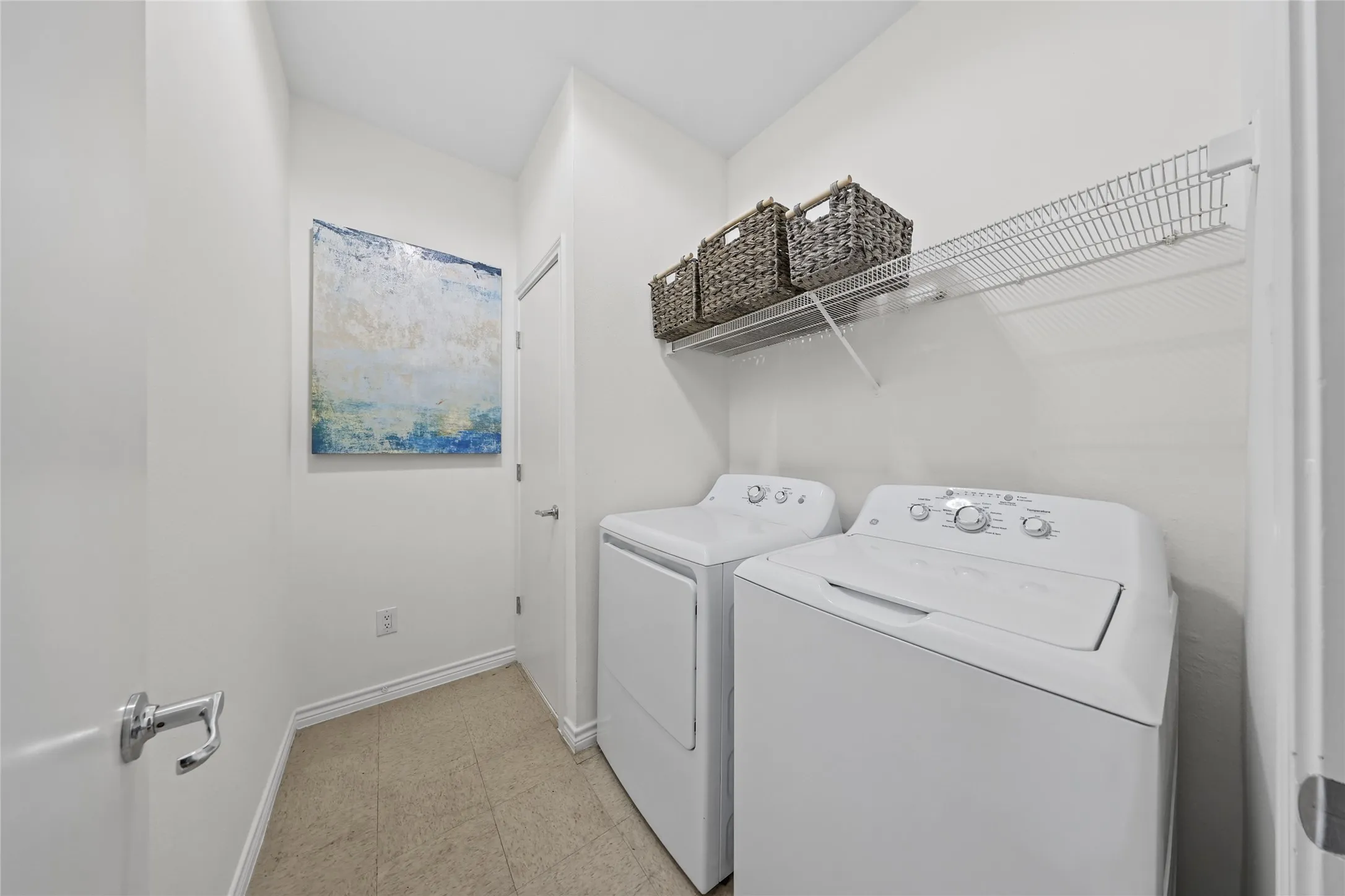 Dedicated laundry room featuring side-by-side washer and dryer with white finishes, wire shelving for storage, neutral tile flooring, and soft overhead lighting
