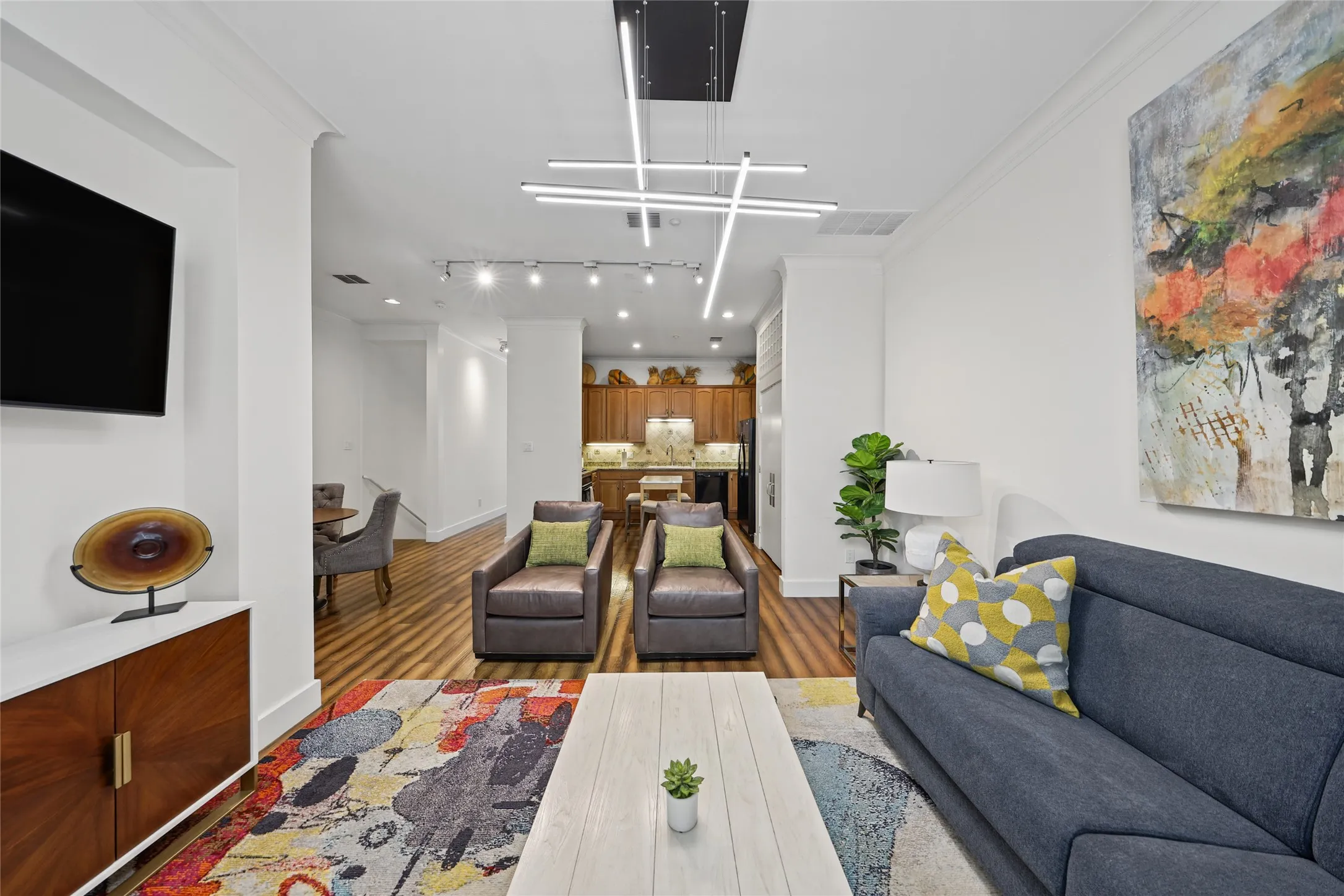 Modern open-concept living space featuring hardwood flooring, recessed and linear ceiling lighting, a gray upholstered sofa with accent pillows, a wood-finish media console with mounted flat-screen television, and a colorful area rug anchoring the seating arrangement
