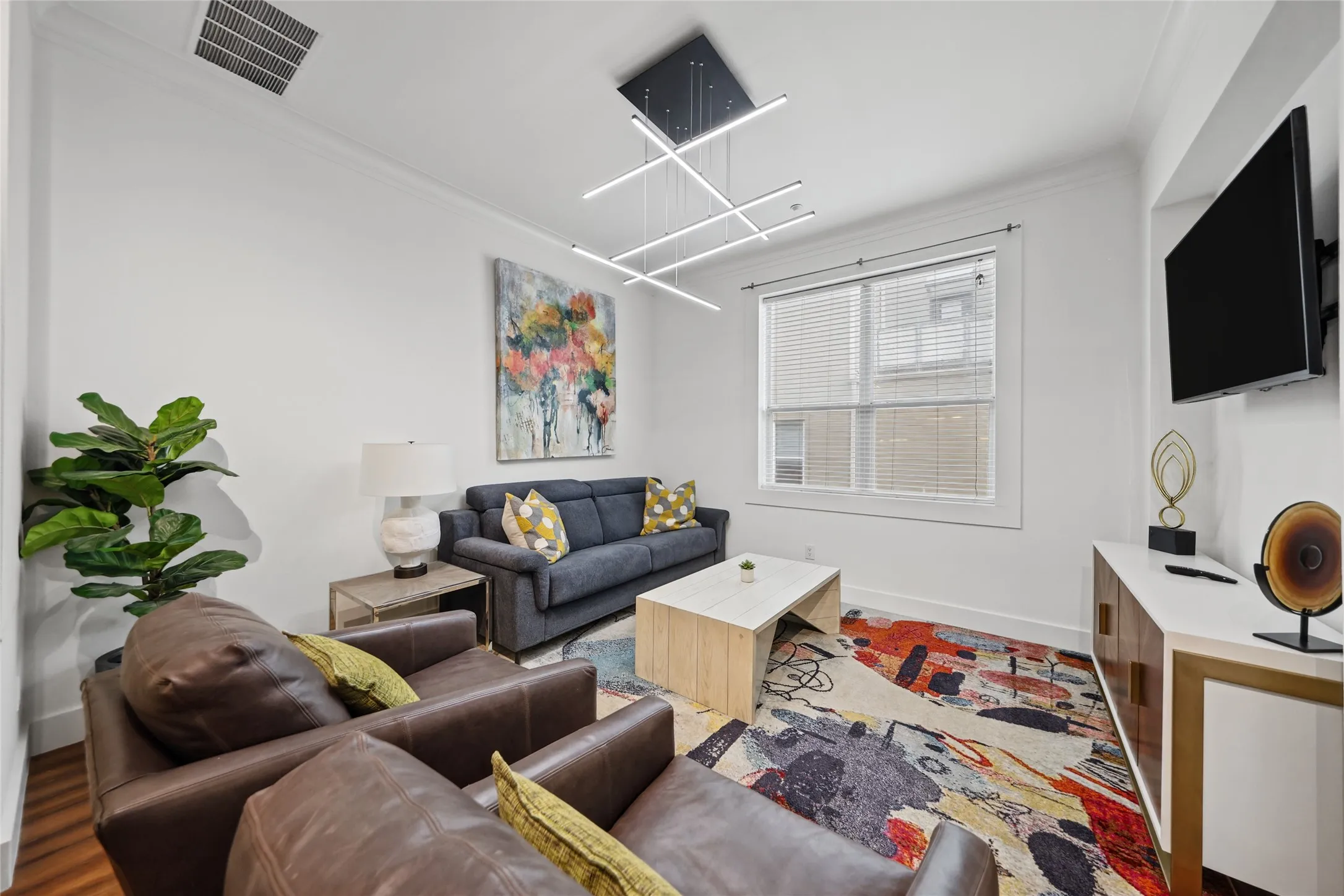 White walls complemented by crown molding, wood-finish flooring, modern linear pendant lighting, and a built-in media console with gold accents