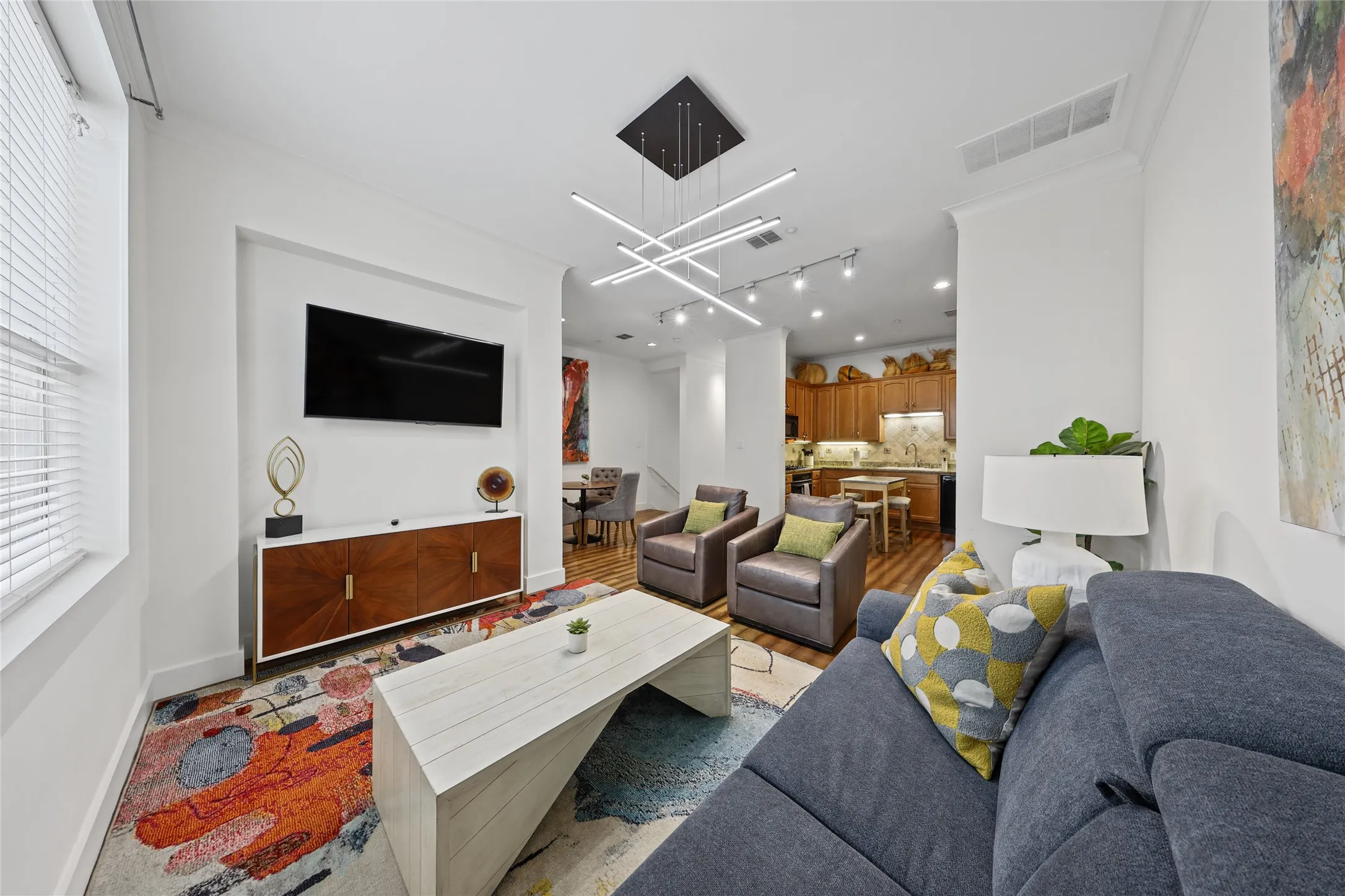 Open-concept living space featuring a wall-mounted flat-screen television, wood-finish cabinetry with storage, modern linear ceiling fixture, recessed lighting, and a wood-finish kitchen with integrated appliances visible in the background