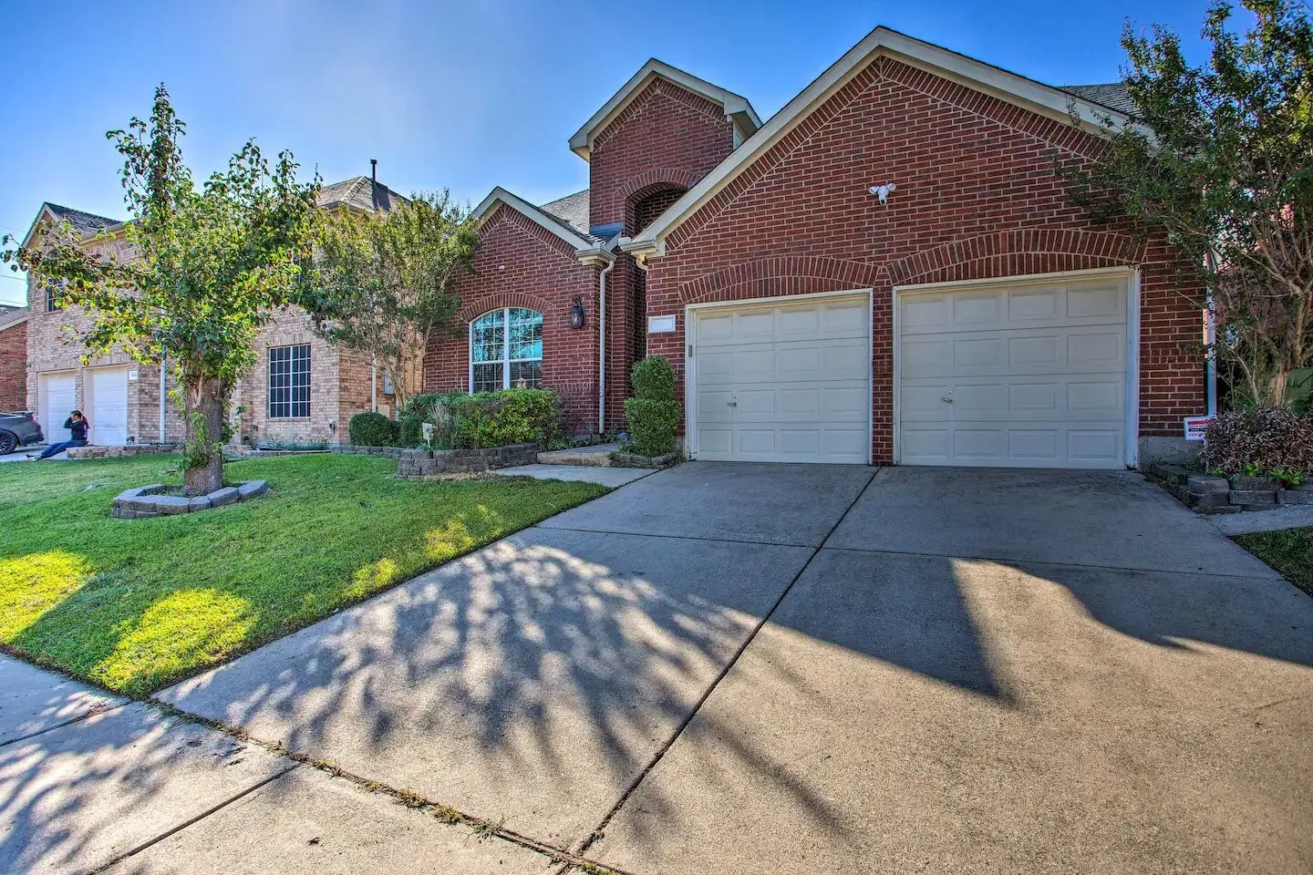 Brick exterior with arched window details, two-car garage, concrete driveway, and front lawn with established landscaping