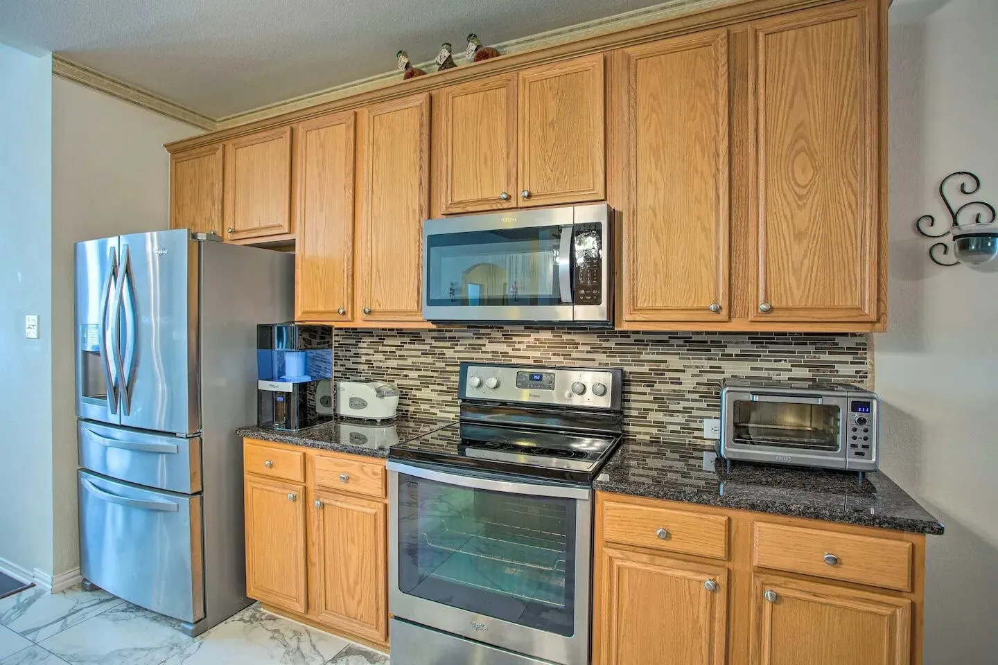 Full-height wood-finish cabinetry, black stone-finish countertops, stainless steel appliances, mosaic tile backsplash, and marble-look flooring