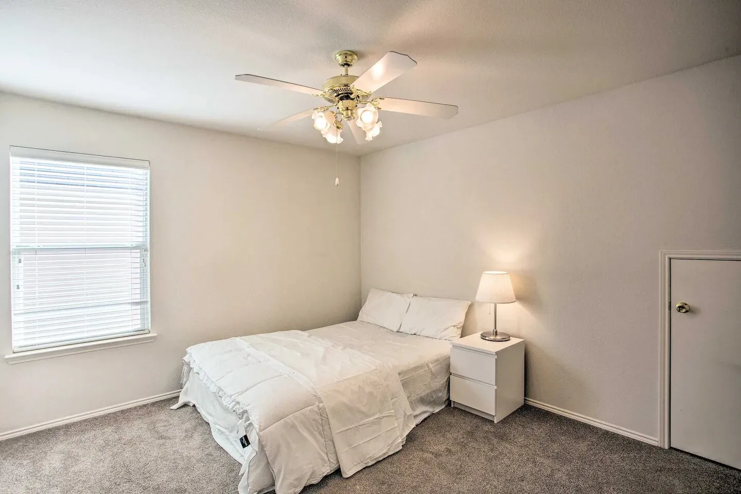 Textured carpeting, a single window with blinds, a ceiling fan with integrated lighting, and a white interior door with a brass-finish knob