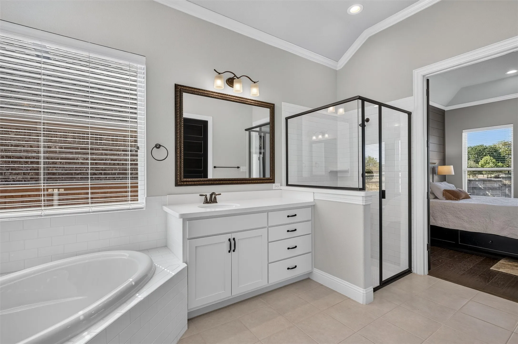 White shaker vanity with quartz countertop, walk-in shower with black frame, soaking tub with white tile surround, and vaulted ceiling