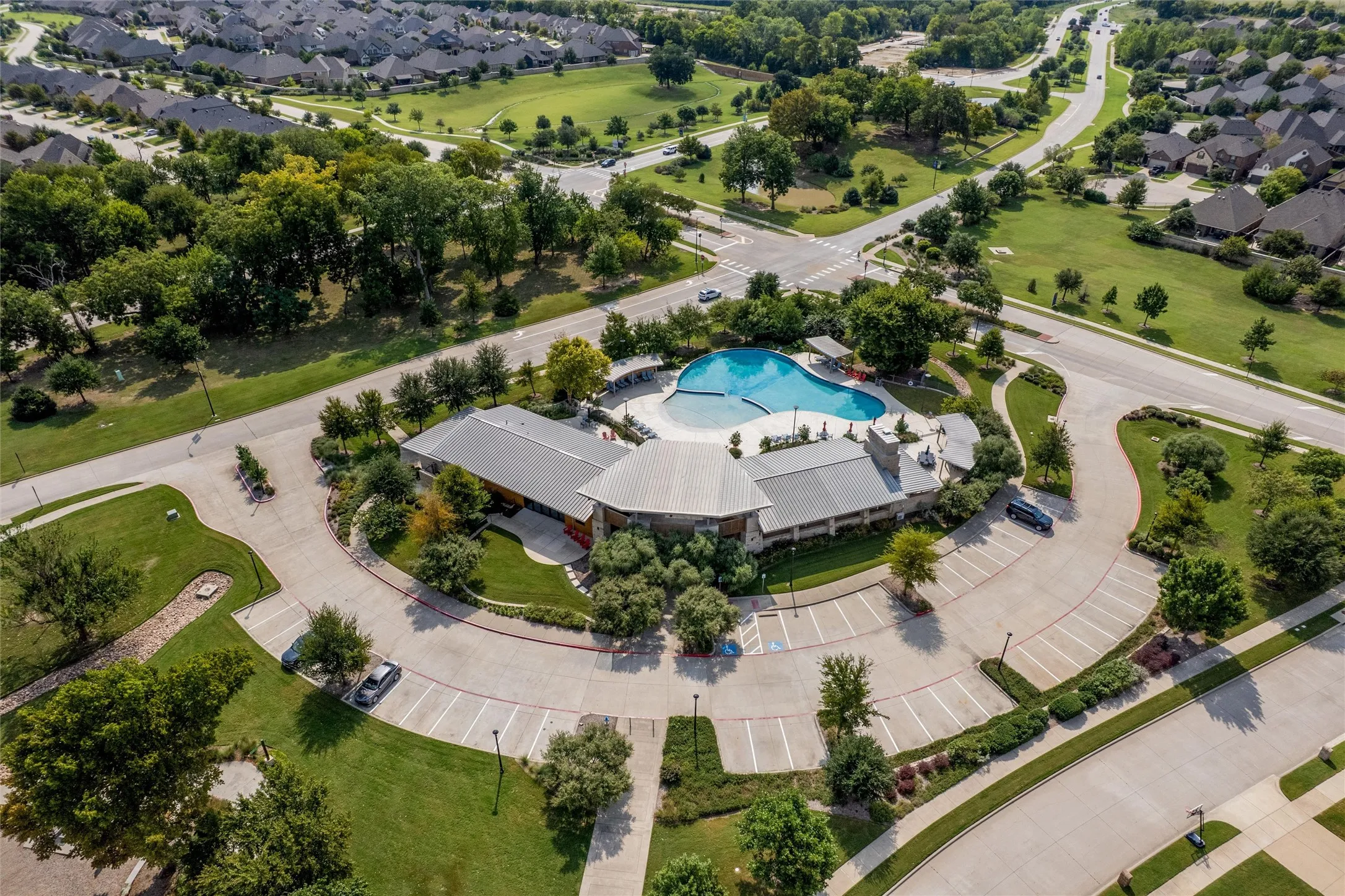 Community clubhouse featuring a large swimming pool, expansive parking, and surrounding green spaces with mature trees