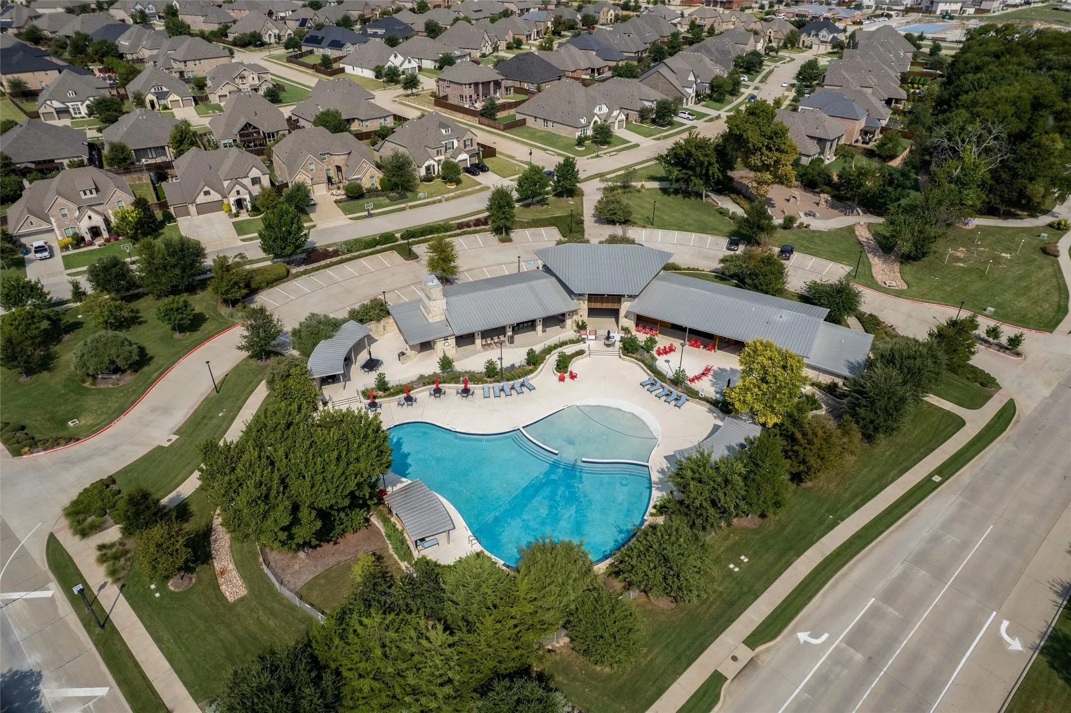 Expansive community pool with curved edges, adjacent clubhouse structures with gabled roofs, surrounding green spaces, and paved parking areas