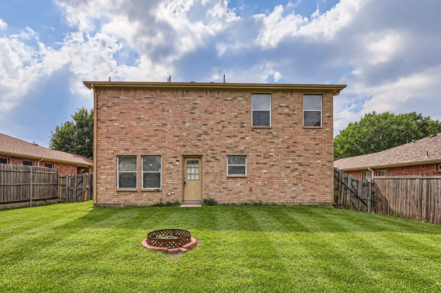 Brick exterior construction with multiple windows, rear entry door, fenced perimeter, and an in-ground fire pit