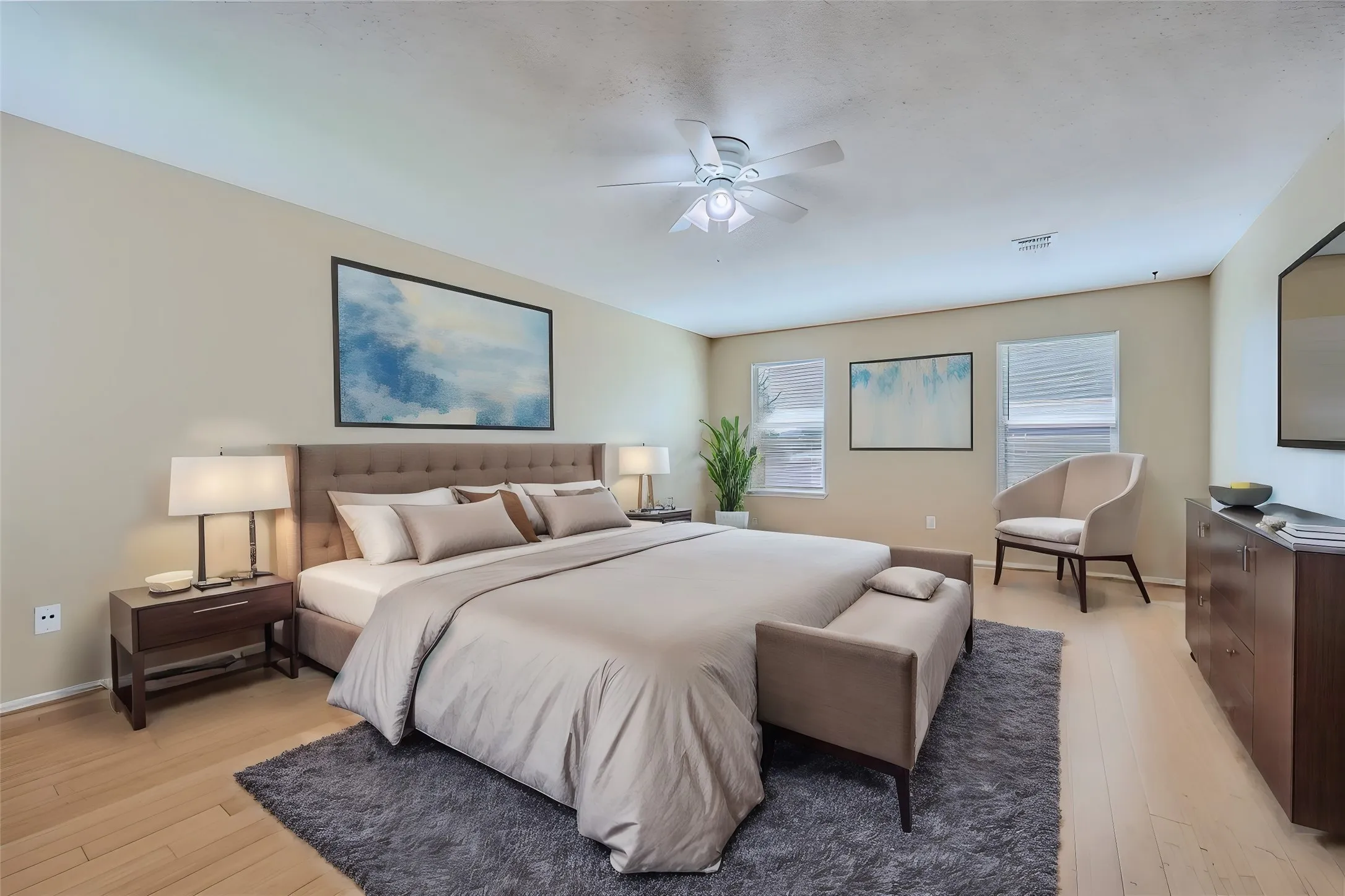 Light wood-finish flooring, tufted headboard, multiple windows with blinds, and ceiling fan with integrated lighting