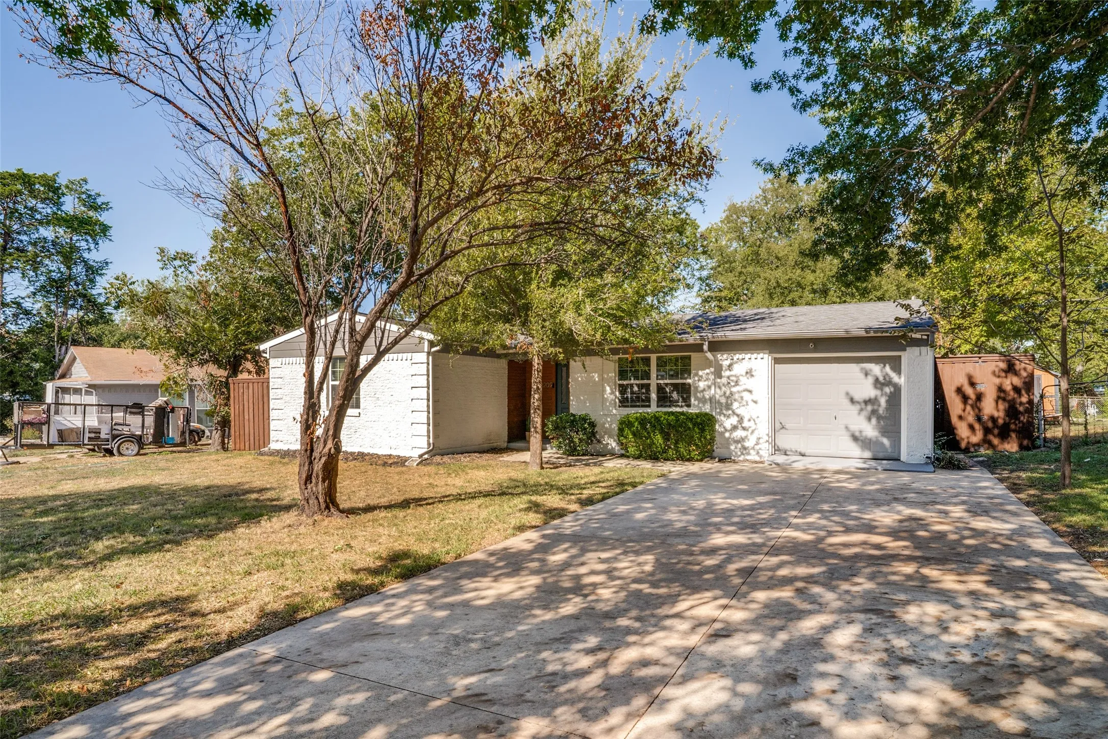 White painted brick exterior with a gray garage door, a concrete driveway, and established trees