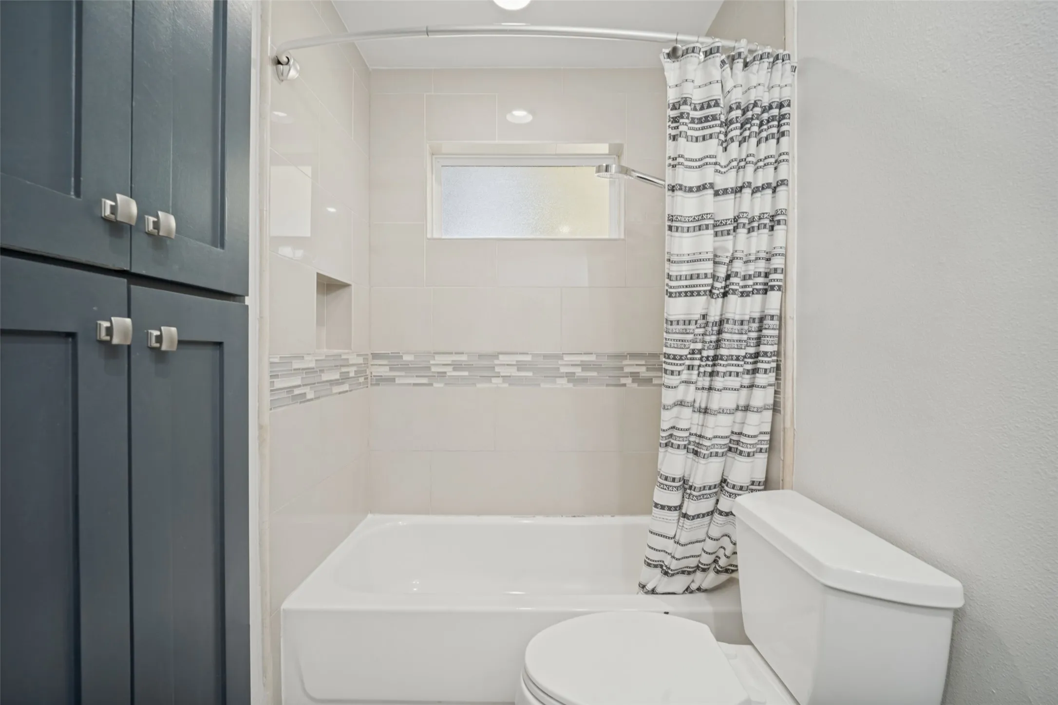 Built-in cabinetry with square pulls, fully tiled shower with accent band, recessed shower niche, frosted window, and recessed lighting