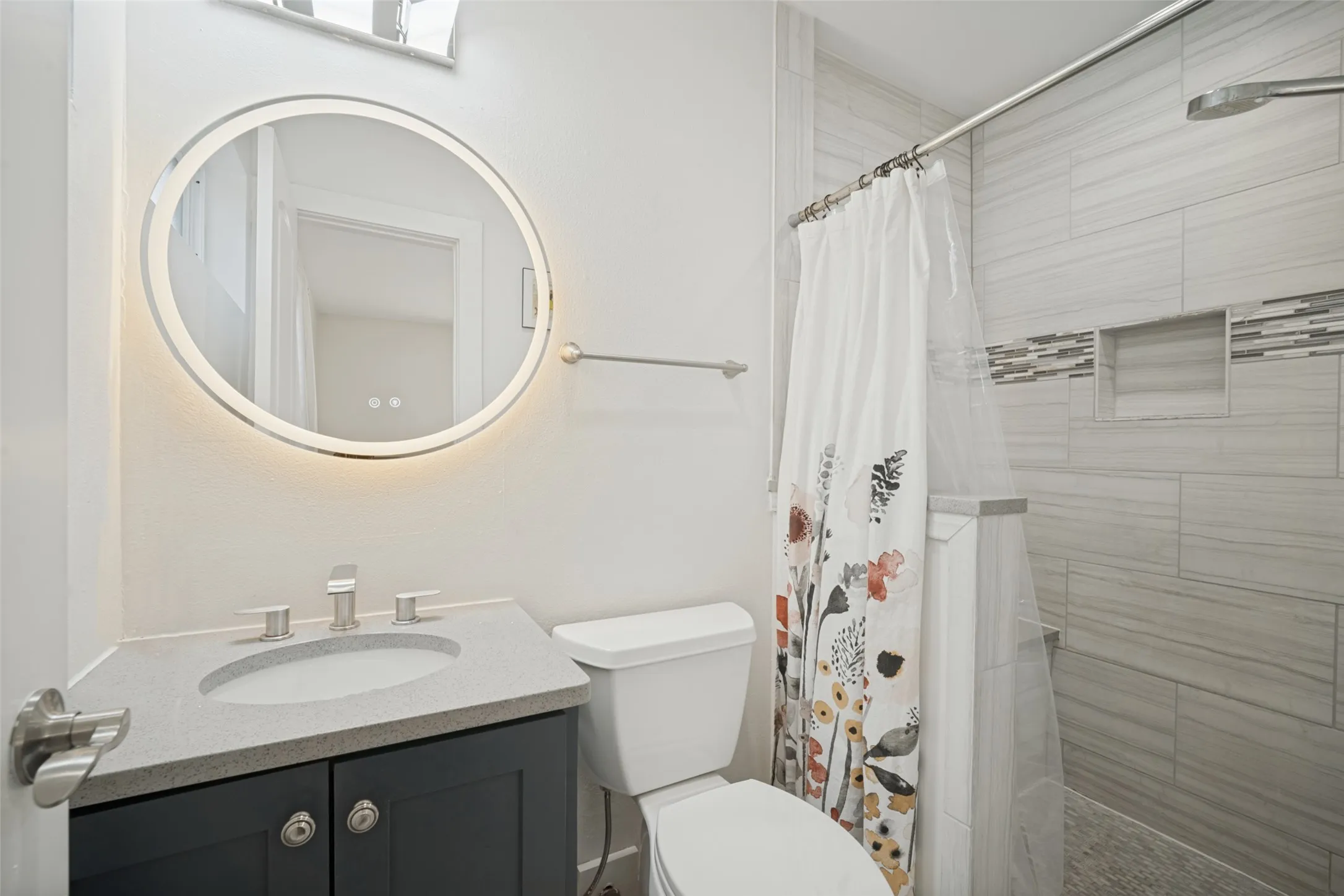 Bathroom featuring a round LED-backlit mirror, dark-finish vanity with an integrated sink, and a tiled shower with a mosaic accent band and built-in niche