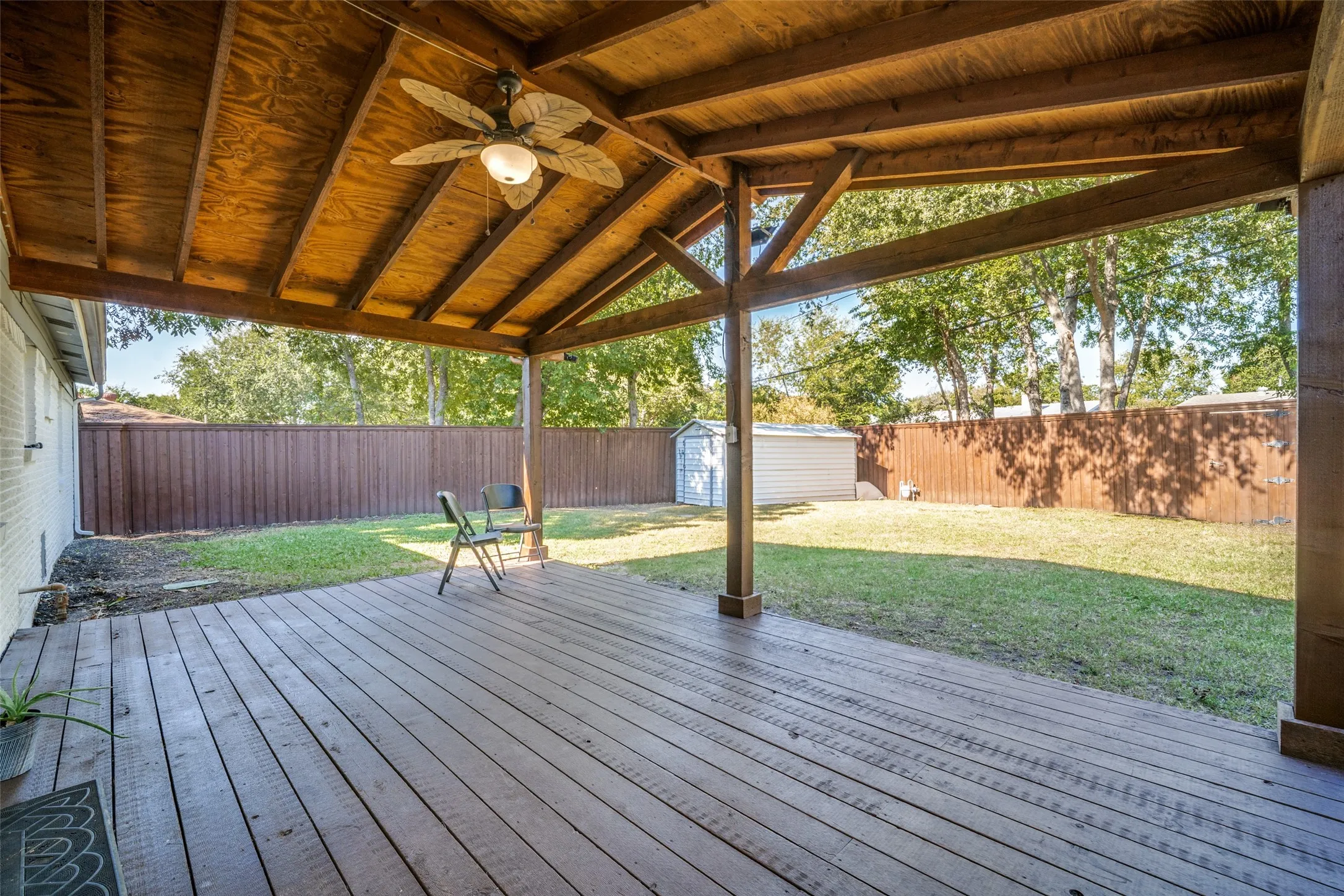 Expansive covered wood deck with a ceiling fan, overlooking a fenced backyard with a detached storage shed