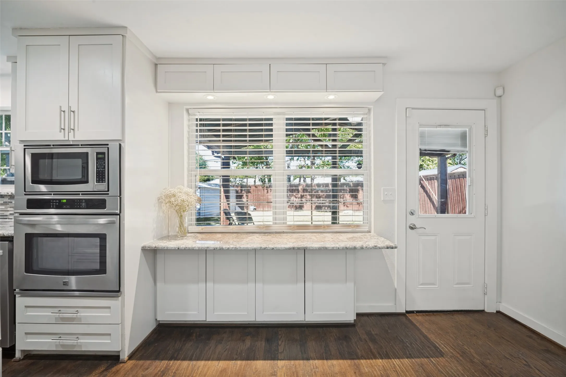Integrated stainless steel microwave and oven, white cabinetry, light-toned countertop, wood-finish flooring, and a glass-paneled exterior door