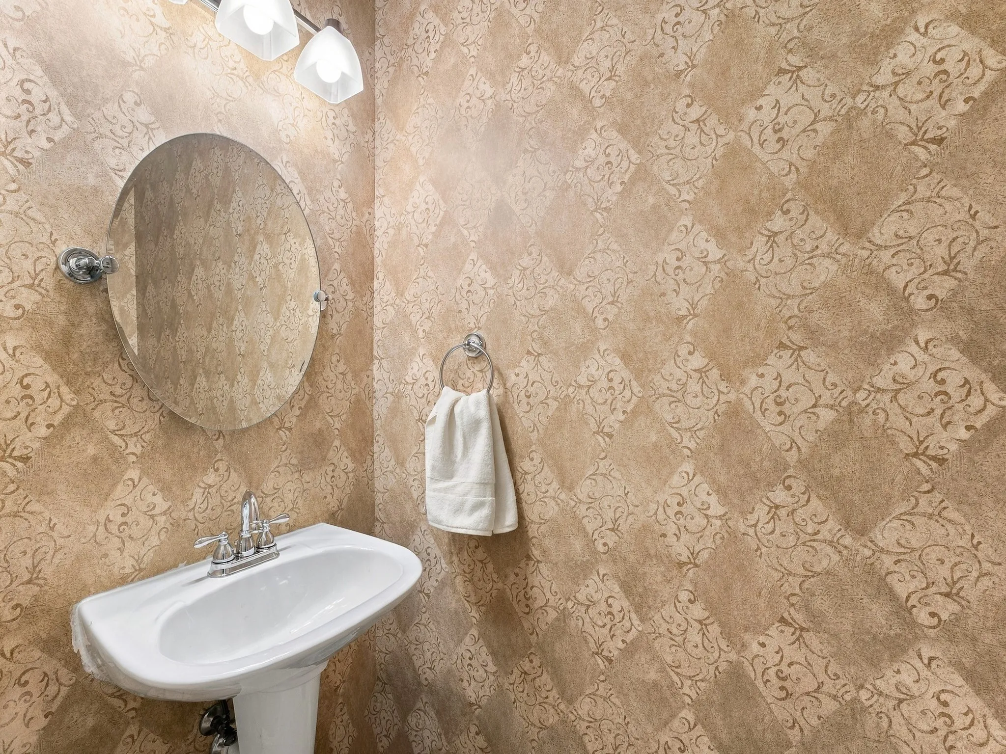 Pedestal sink with chrome faucet, oval wall-mounted mirror, and decorative wall covering