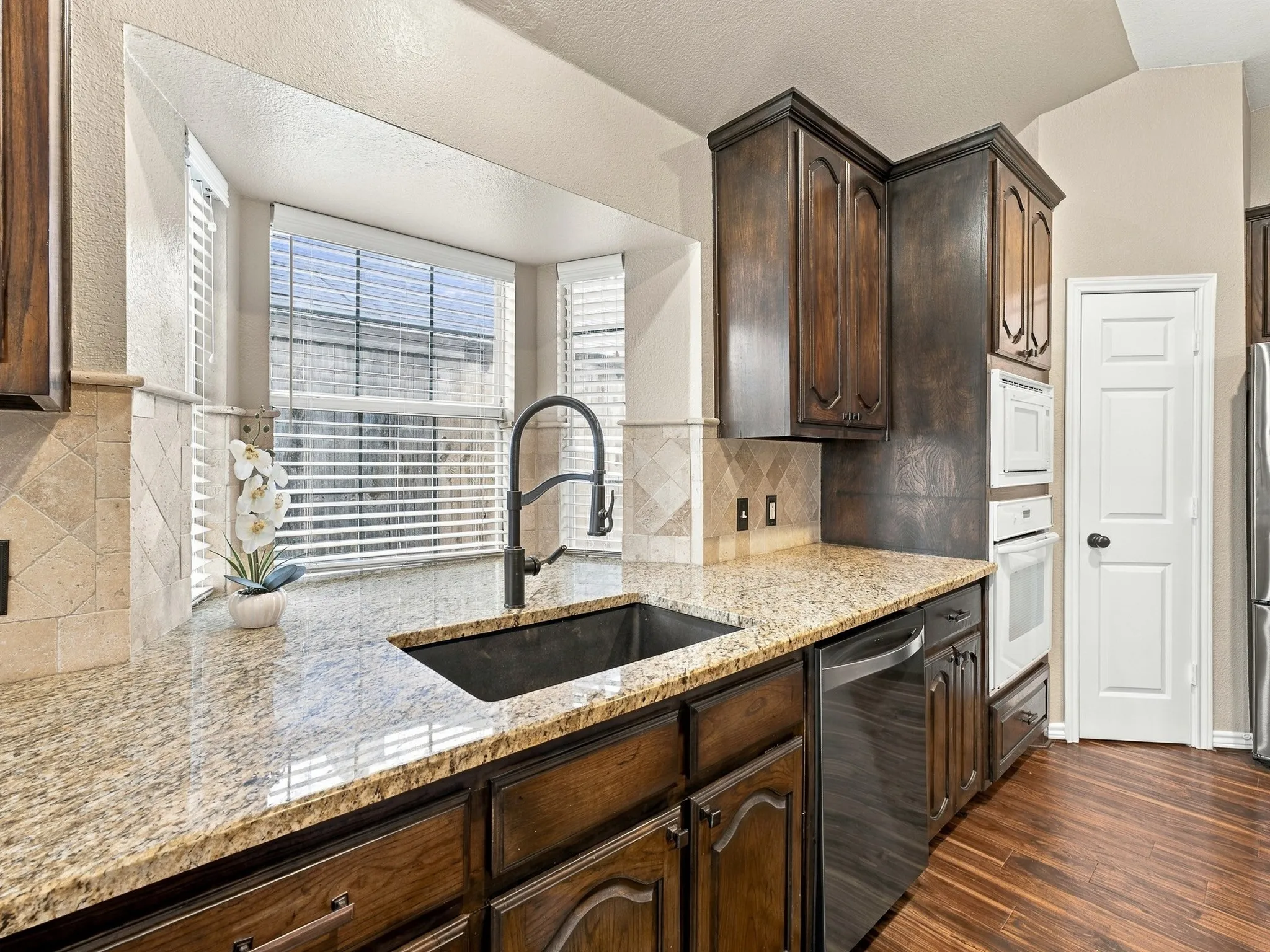 Kitchen featuring wood-finish flooring, granite countertops, dark wood cabinetry, a built-in oven, and a large sink with a gooseneck faucet