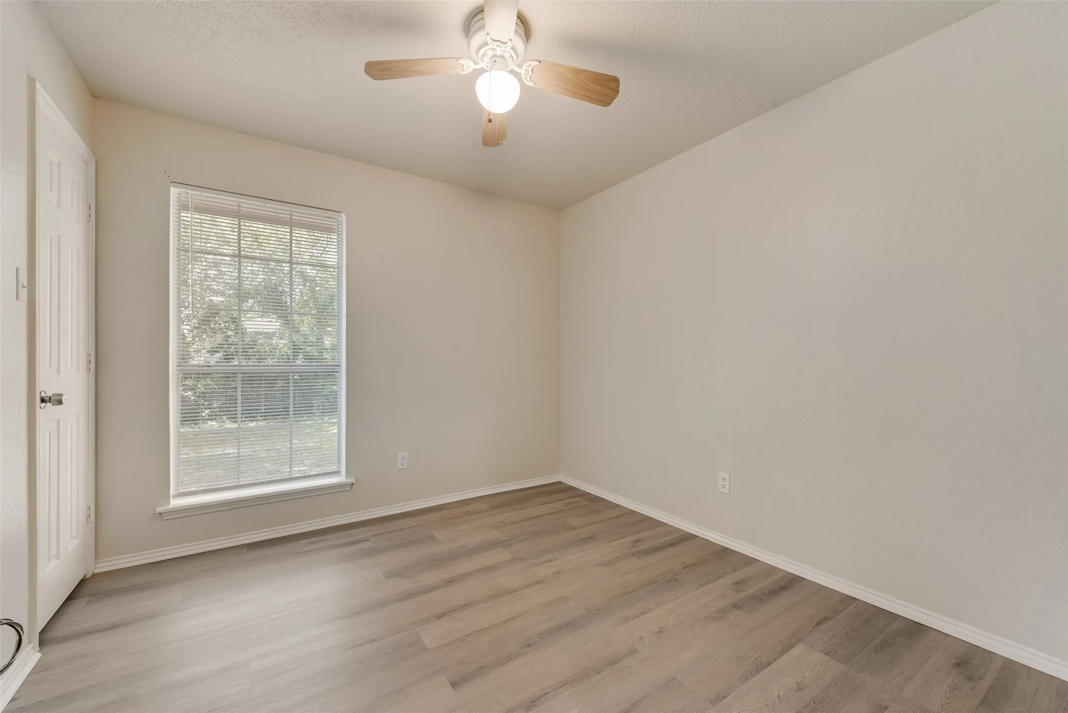 Wood-finish flooring, single window with blinds, ceiling fan with integrated lighting, and a white paneled door