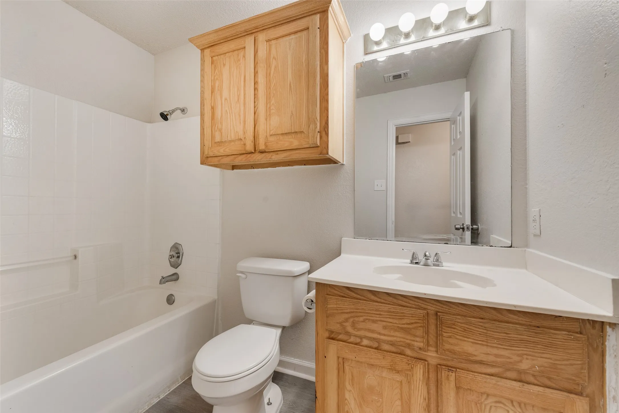 White tub/shower combination, wood-finish vanity with an integrated sink, and overhead wood-finish cabinetry