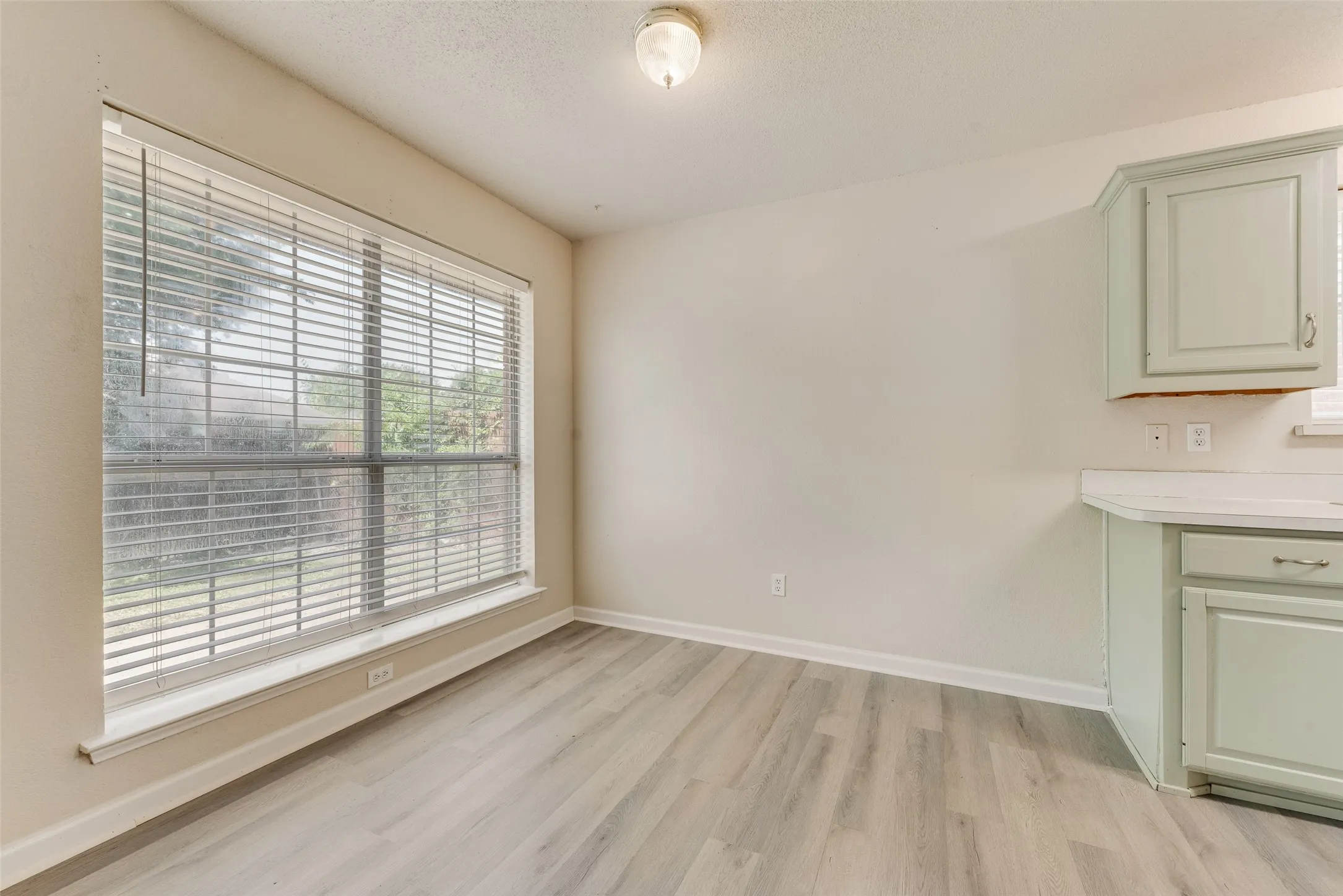 Light wood-finish flooring, a large window with horizontal blinds, built-in cabinetry with hardware, and solid surface countertops