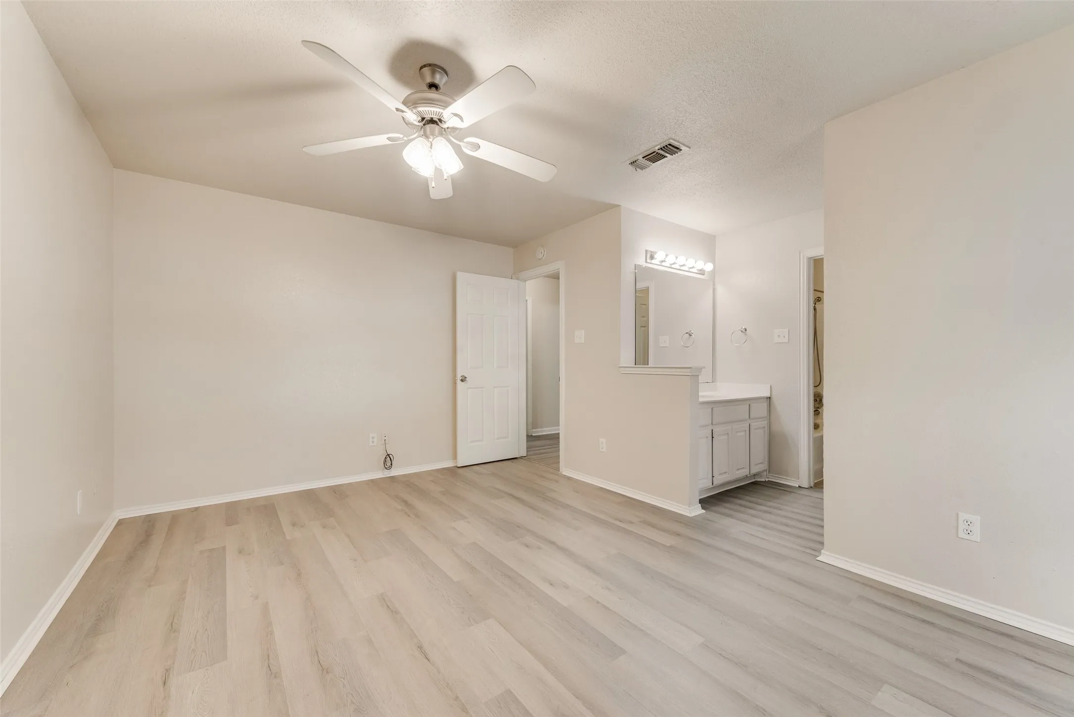 Spacious room with light wood-finish flooring, ceiling fan, and access to a vanity area with built-in cabinetry, a rectangular mirror, and vanity lighting