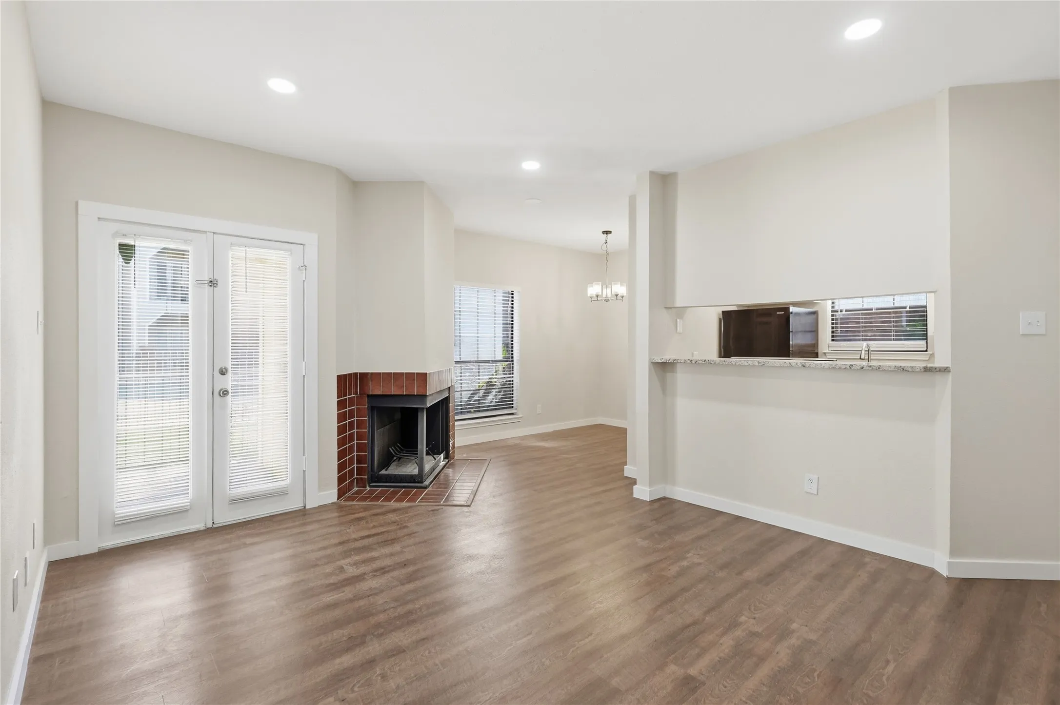 Wood-finish flooring, recessed lighting, brick-clad fireplace, double French doors, and a built-in pass-through window with a granite-style countertop