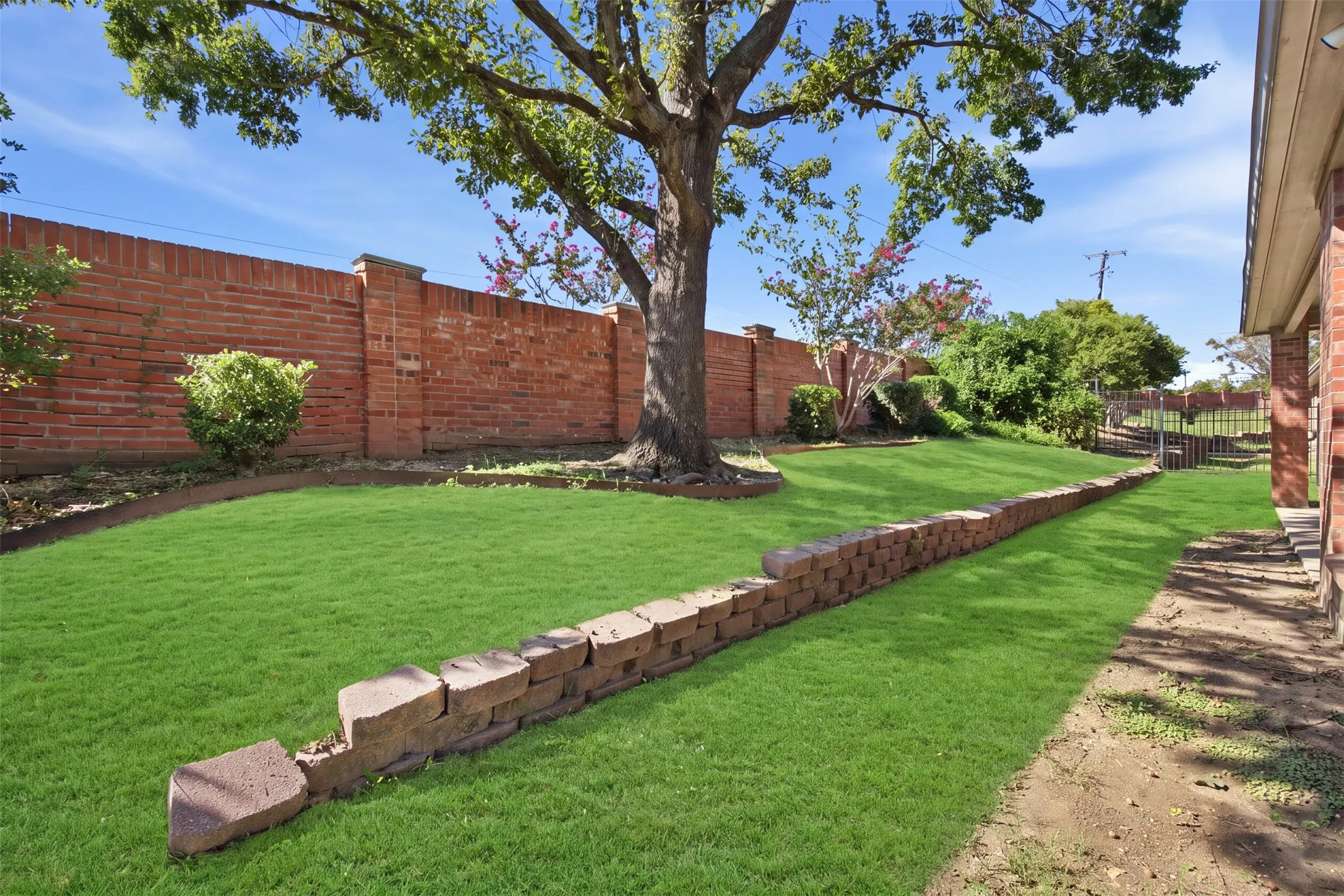 Green lawn with mature shade trees, bordered by a brick privacy wall and a stone retaining wall. Green enhancement to lawn.