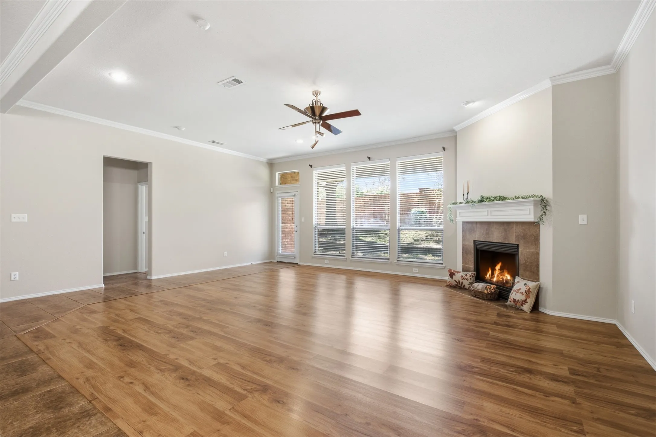 Spacious living area featuring wood-finish flooring, a ceiling fan, a tiled fireplace with a white mantel, and multiple windows with blinds