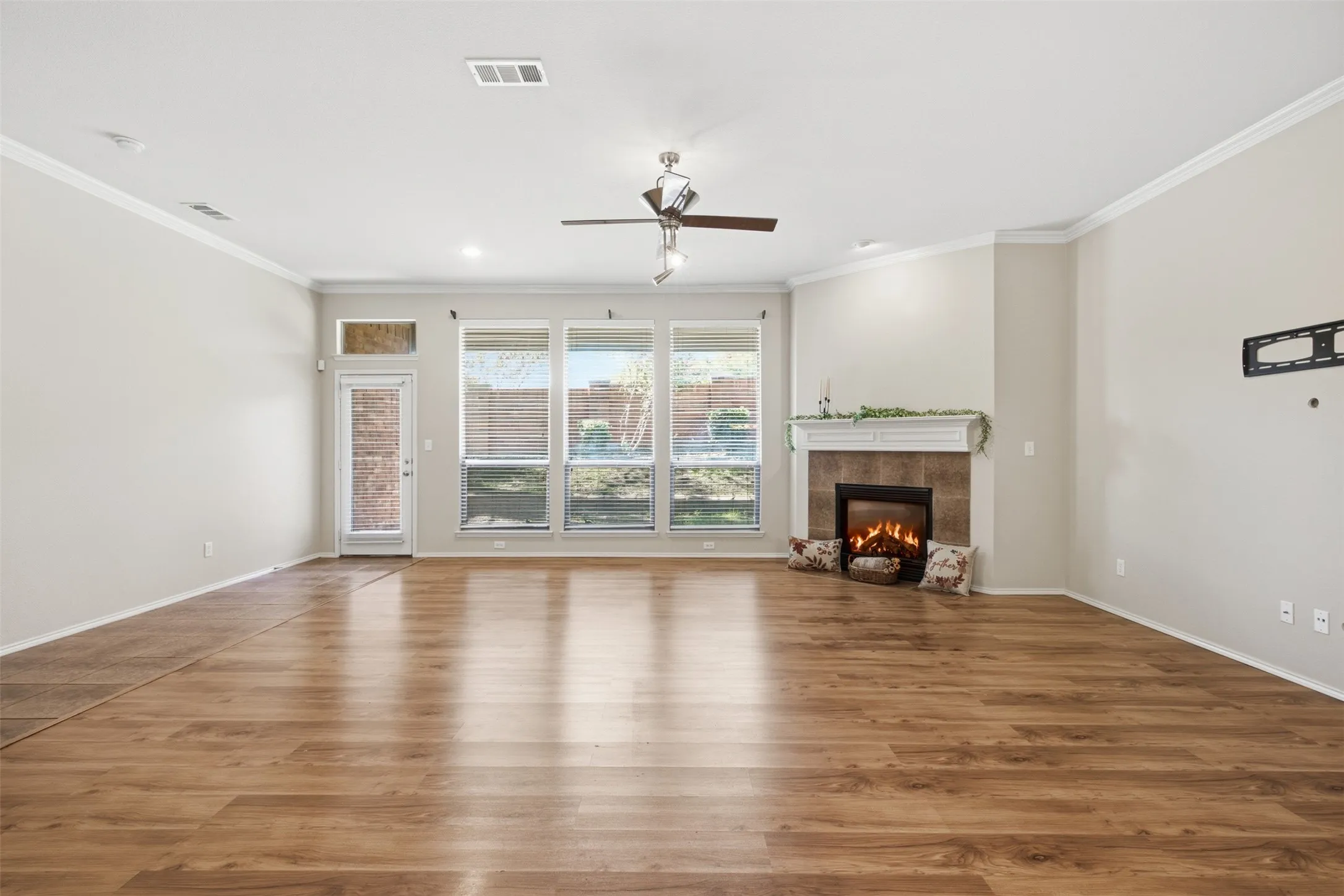 Wood-finish flooring throughout, a tiled fireplace with a white mantel, multiple large windows, a glass-paned door, and a ceiling fan with integrated lighting