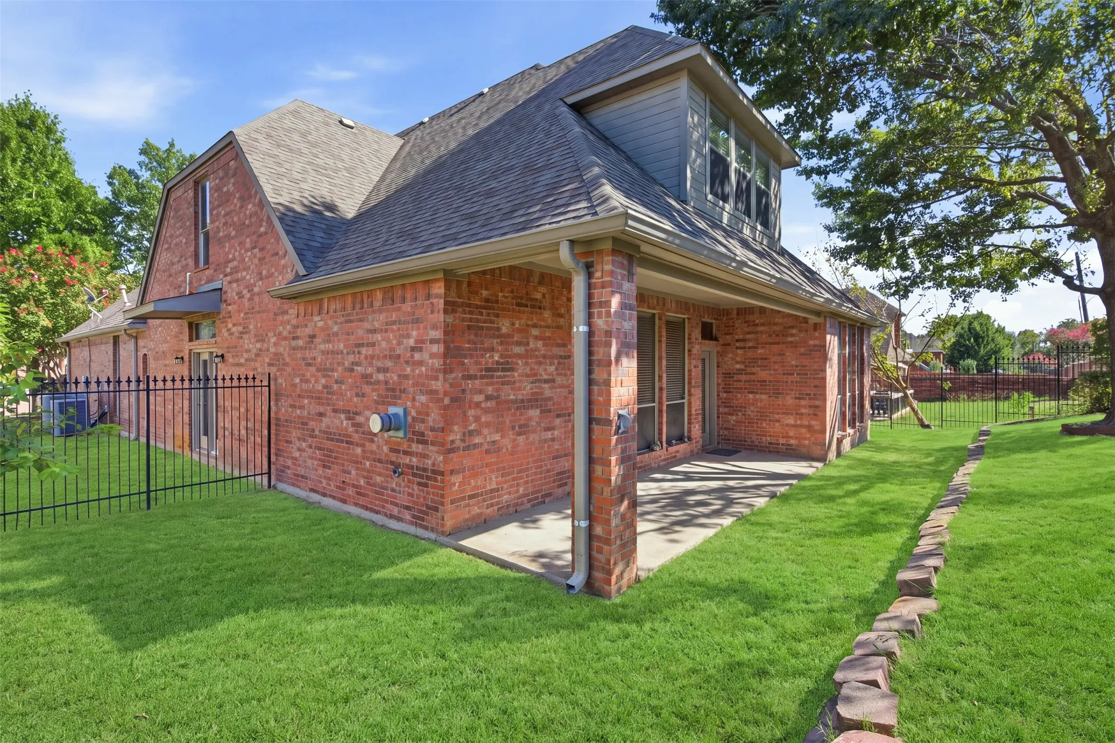 Brick exterior with a covered patio, dormer window, and asphalt shingle roof. Green enhancement to lawn.