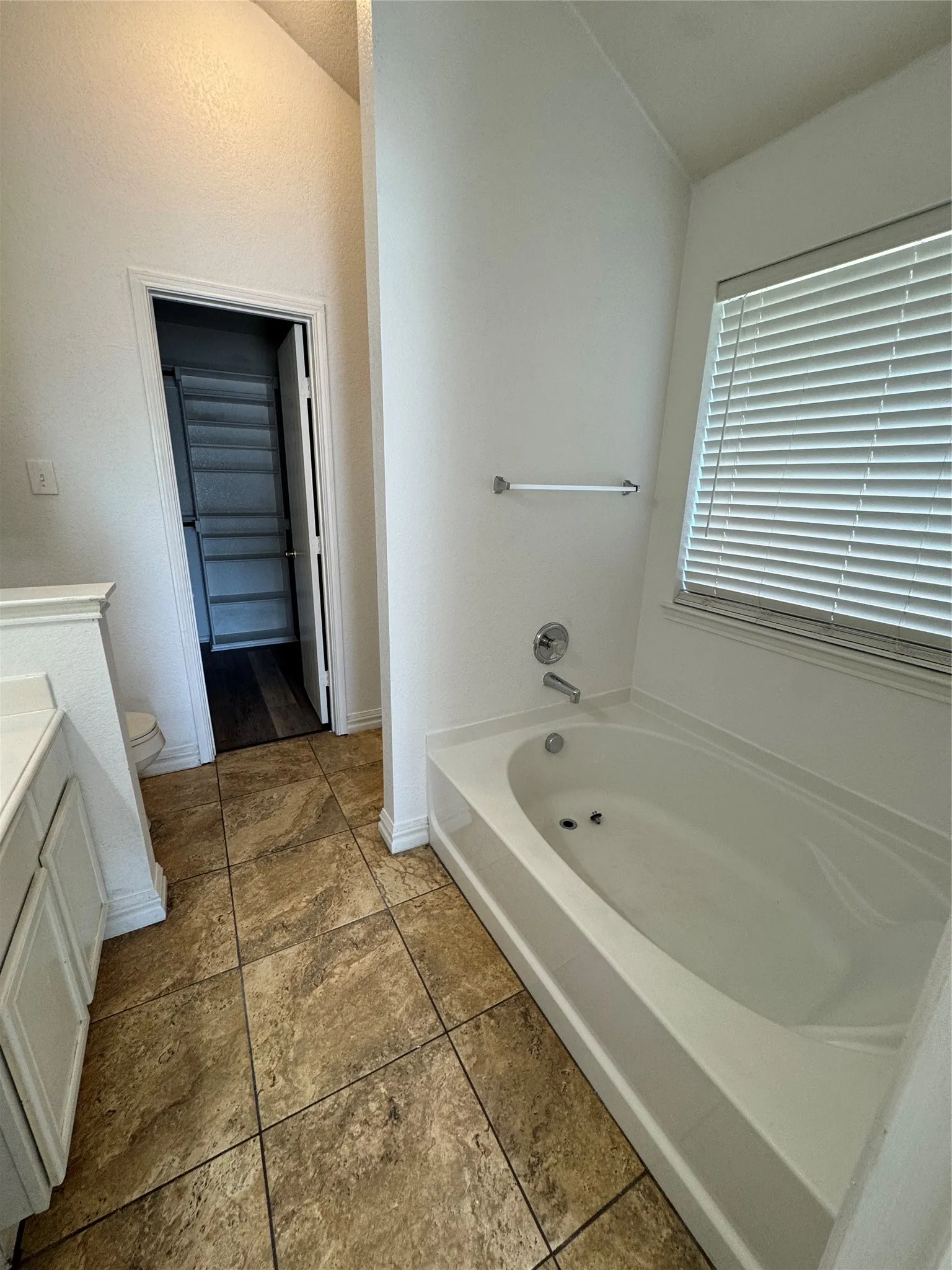 Soaking tub with chrome fixtures, tiled flooring, white vanity with cabinet storage, and a window with blinds