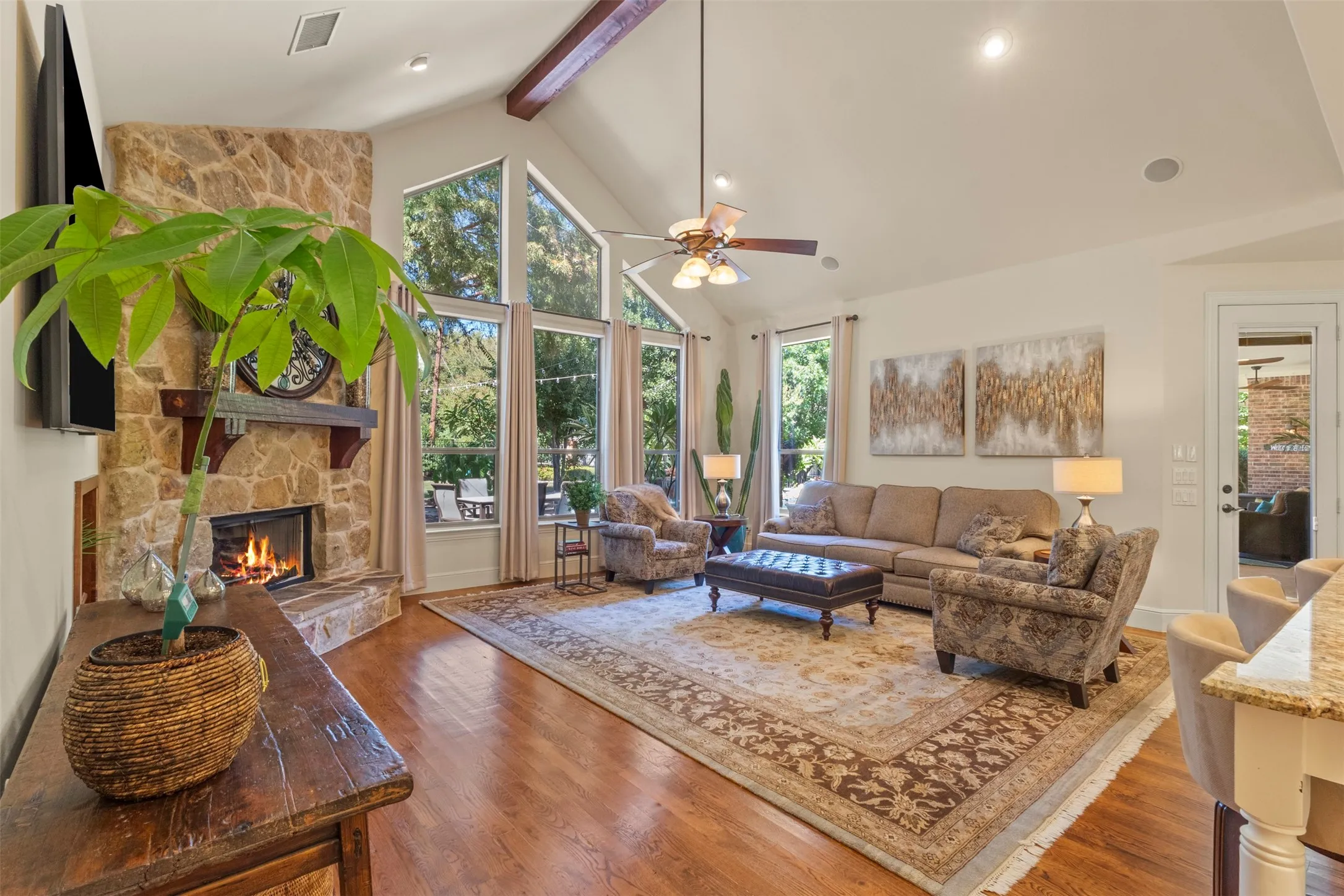 Vaulted ceiling with exposed wood beams, stone fireplace, large picture windows, wood-finish flooring, and recessed lighting