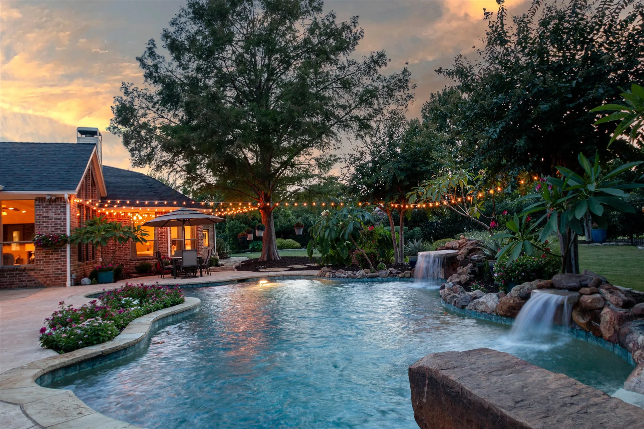 Freeform swimming pool with multiple waterfalls, extensive surrounding hardscaping, and mature landscaping