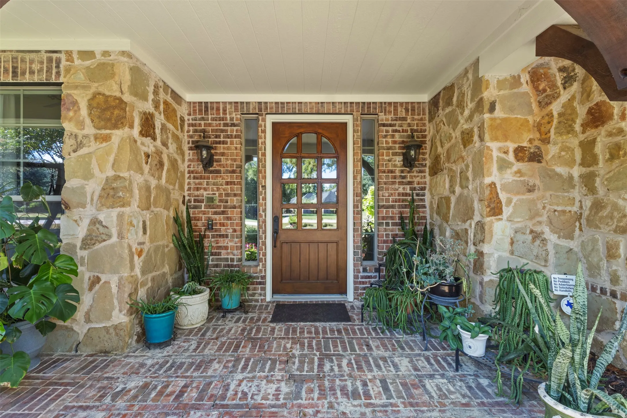 Arched wood-panel entry door, brick-patterned flooring, stone and brick exterior accents, and dual exterior light fixtures