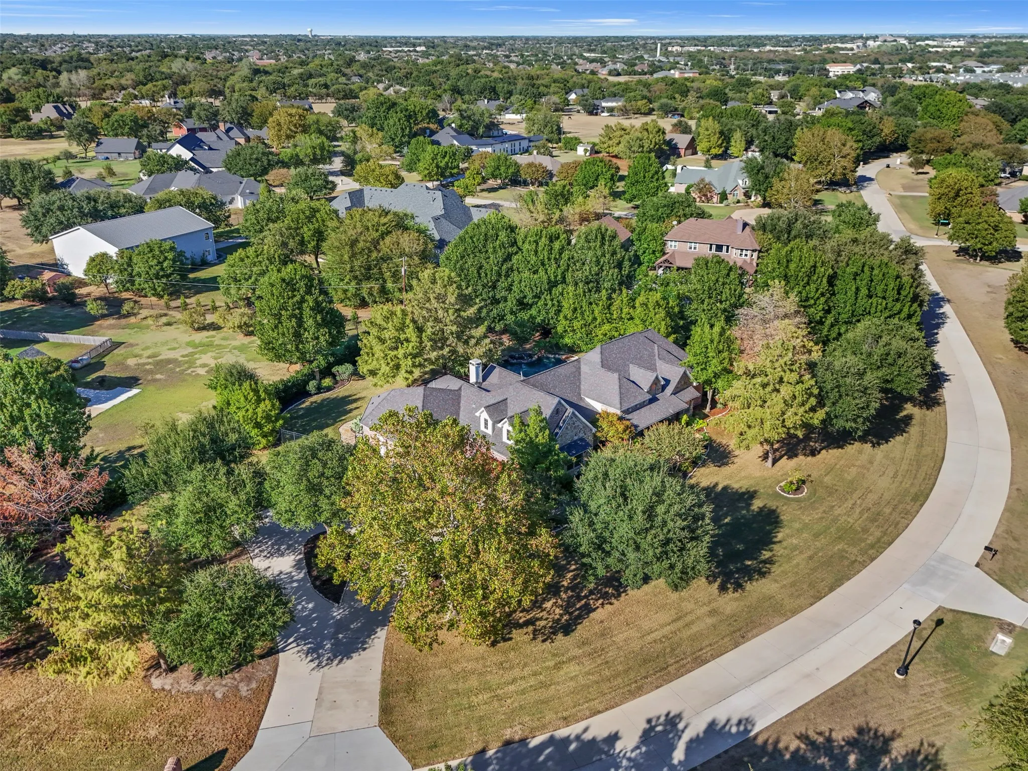 Aerial perspective showcasing a property surrounded by mature trees, a curved concrete driveway, and a shingled roofline