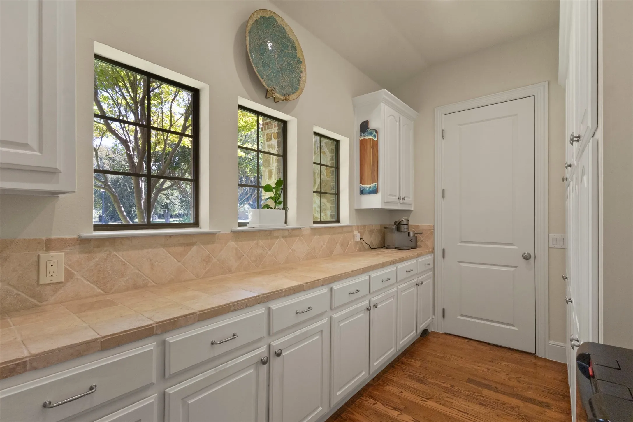 White cabinetry, tile countertops, tile backsplash, wood-finish flooring, and multiple windows