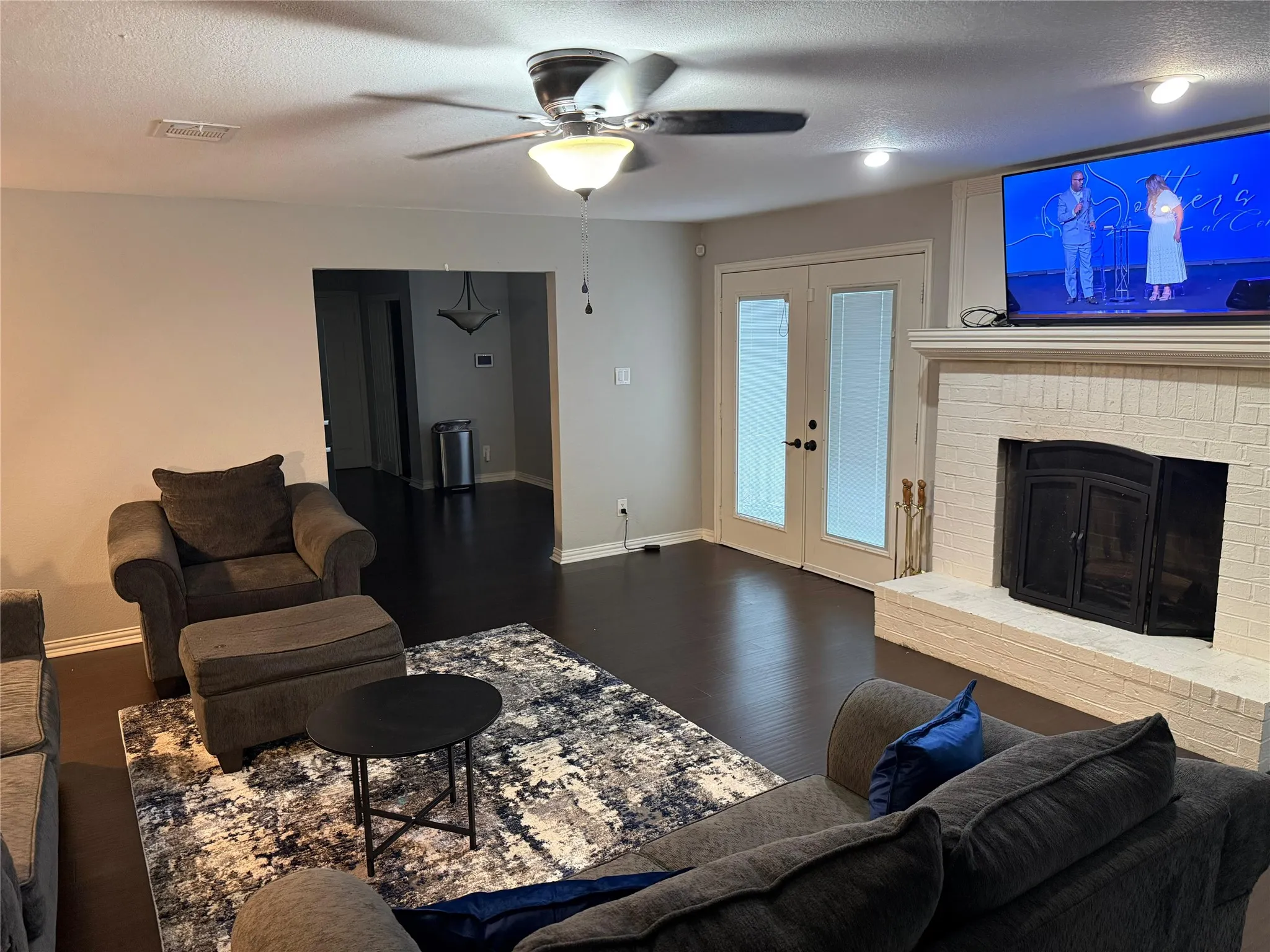 Dark wood-finish flooring, white brick fireplace with mantel, double glass-paneled doors, ceiling fan with integrated lighting, and recessed ceiling lighting