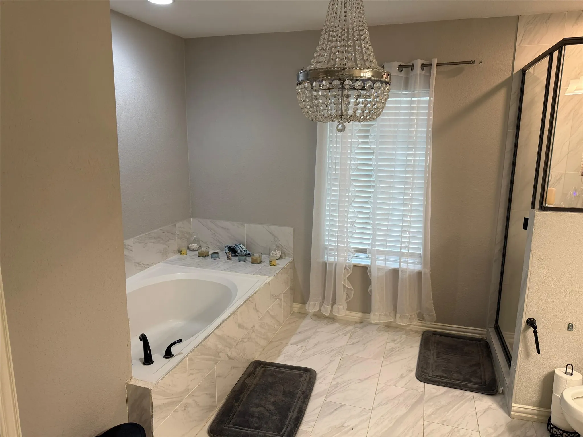 Built-in soaking tub with black fixtures, tiled surround, white window blinds, and crystal chandelier