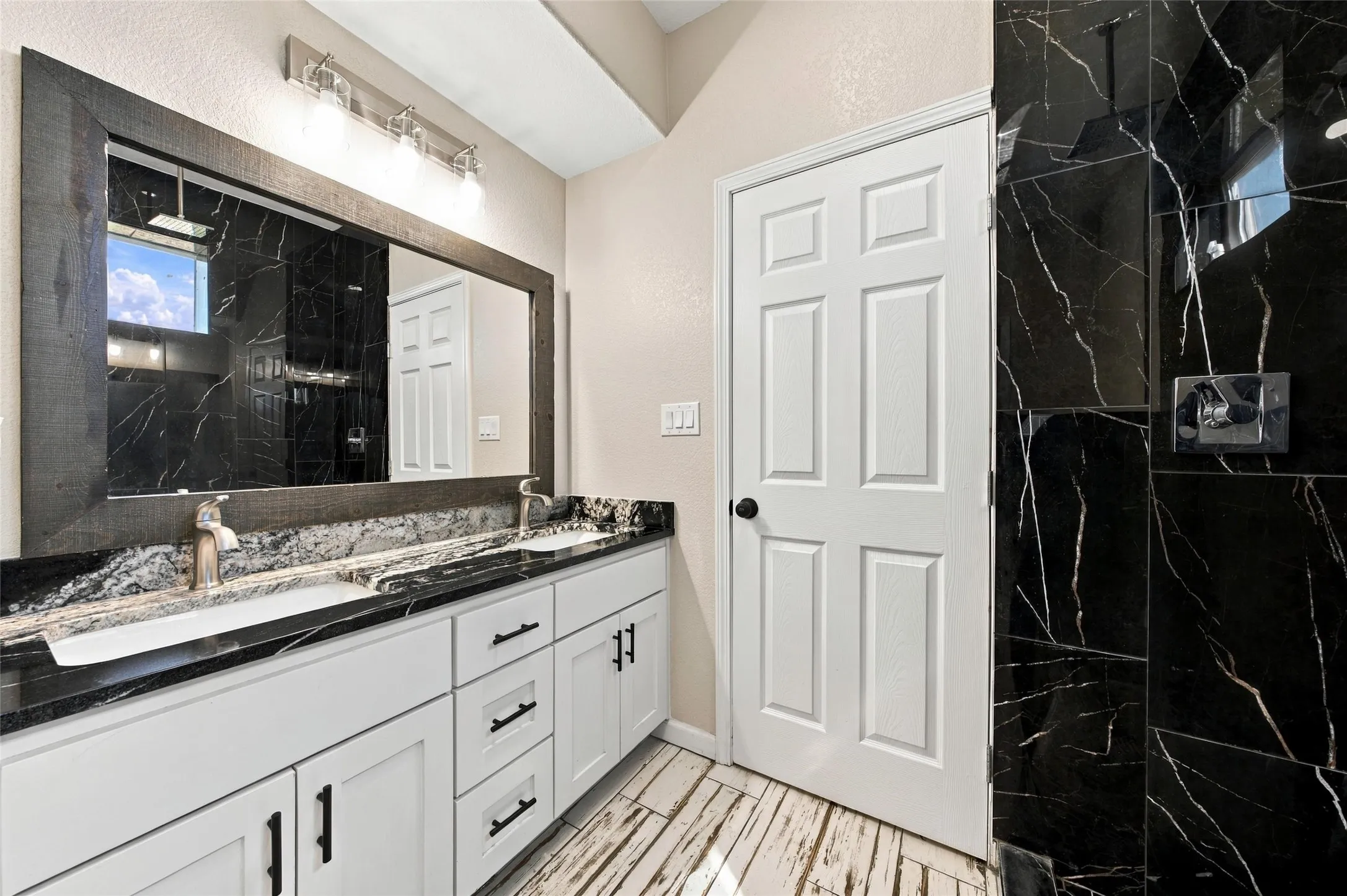 Double vanity with undermount sinks, black and white veined countertop, white shaker cabinetry, and wood-finish flooring