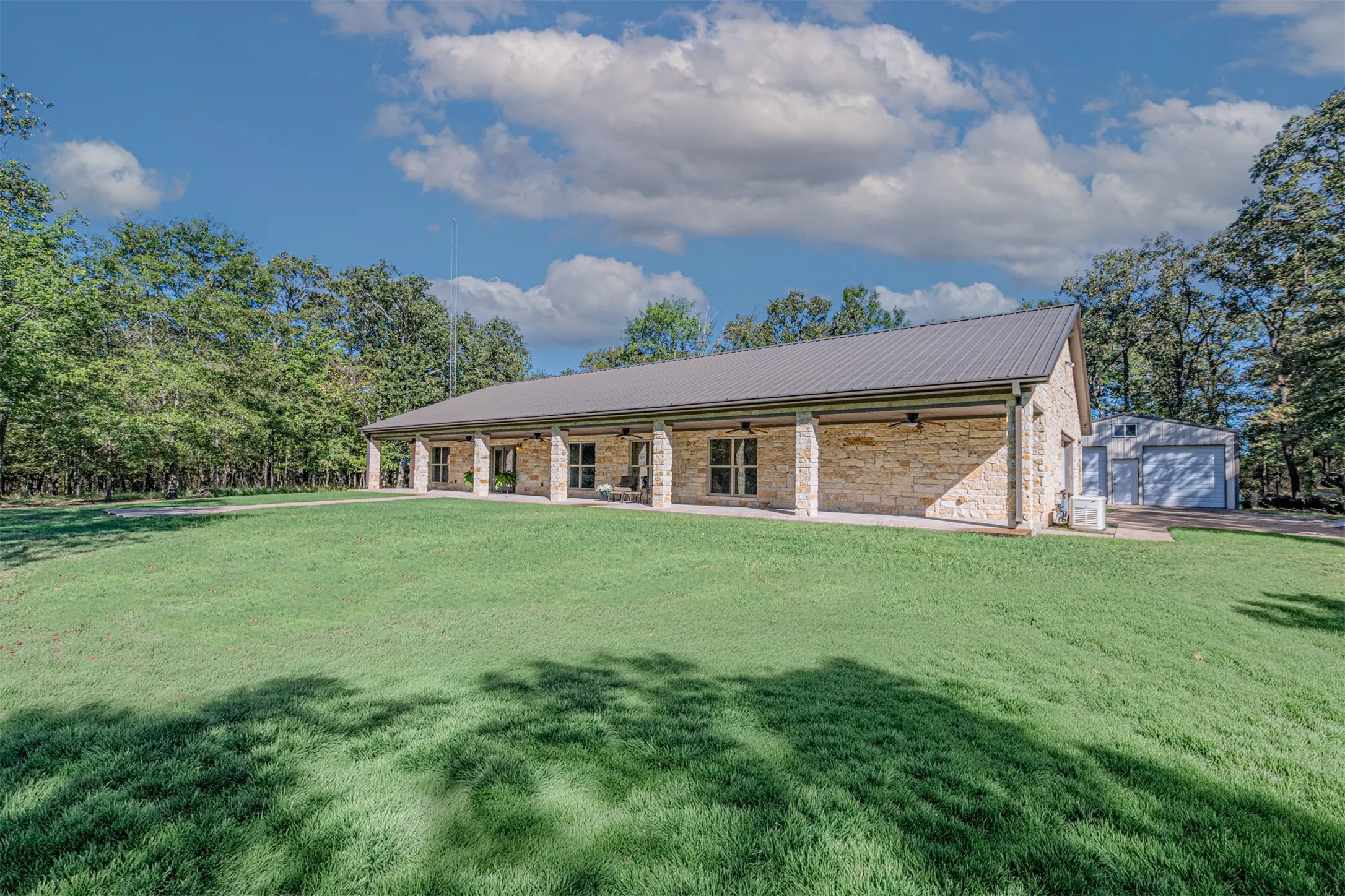 Stone-clad exterior with a metal roof, extended covered porch, multi-panel windows, and a detached garage structure