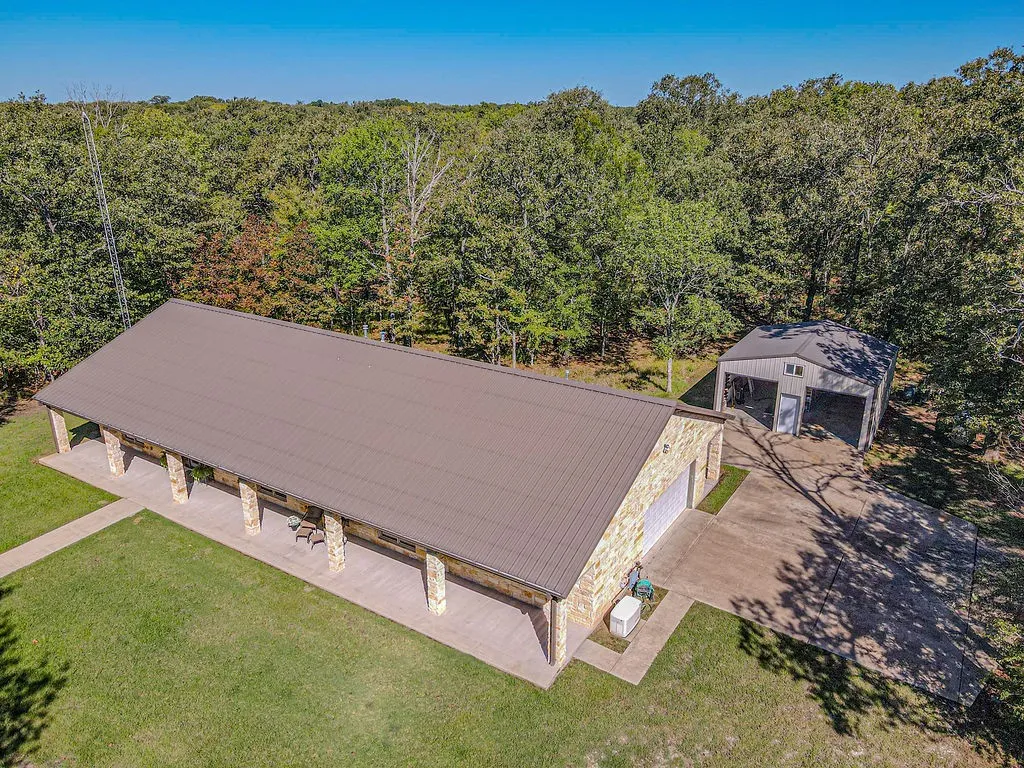Stone-clad exterior, metal roofing, extended covered patio with stone columns, attached two-car garage, and separate detached two-bay outbuilding