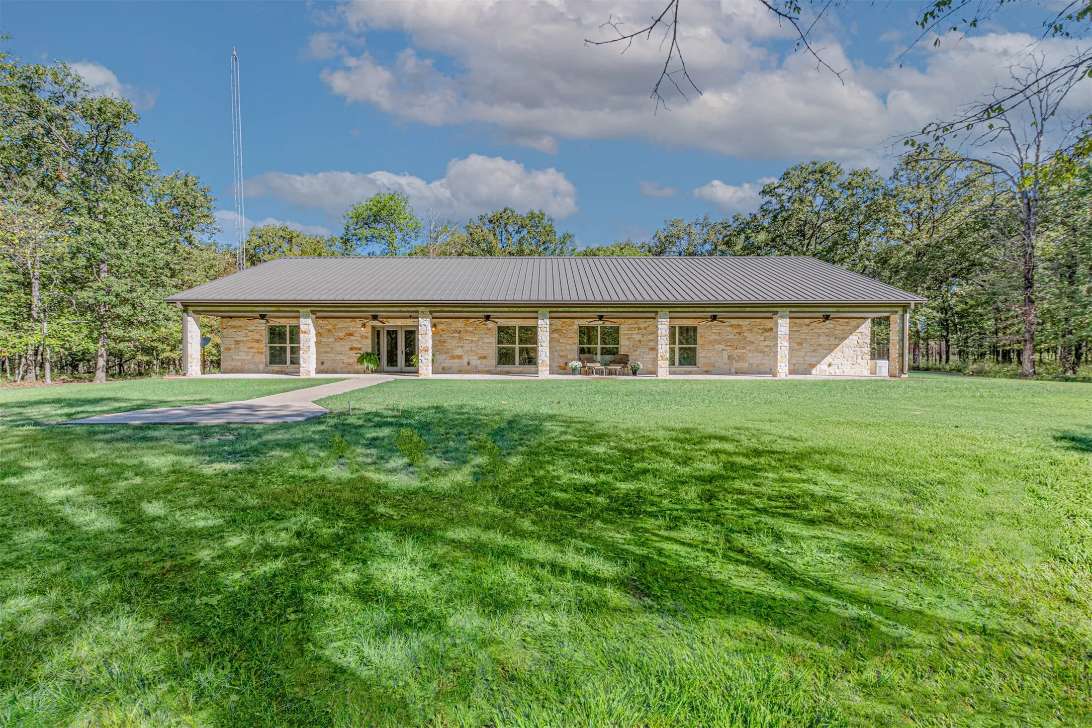 Single-story stone exterior, metal roof, covered front porch, concrete patio, and mature trees