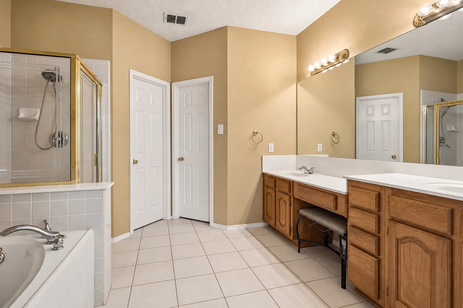 Double vanity with integrated sinks and a dedicated makeup area, large wall-mounted mirror, separate shower enclosure with a glass door, built-in bathtub with tiled surround, and ceramic tile flooring