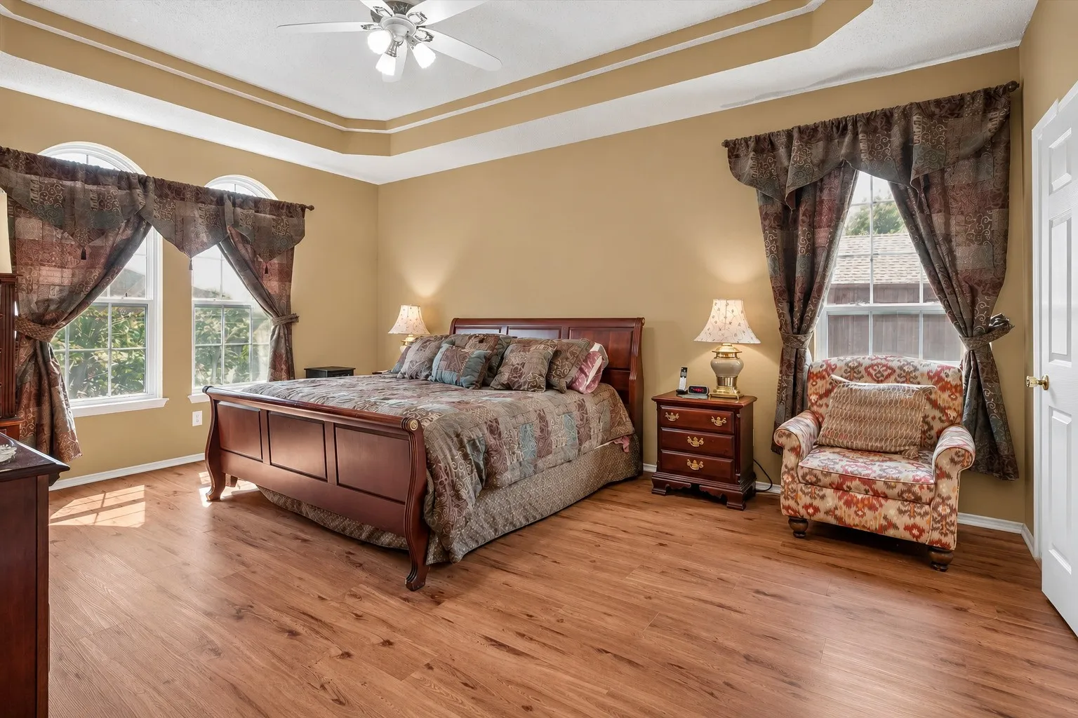 Wood-finish flooring throughout, tray ceiling with crown molding, ceiling fan with integrated lighting, dual arched windows, and a single rectangular window
