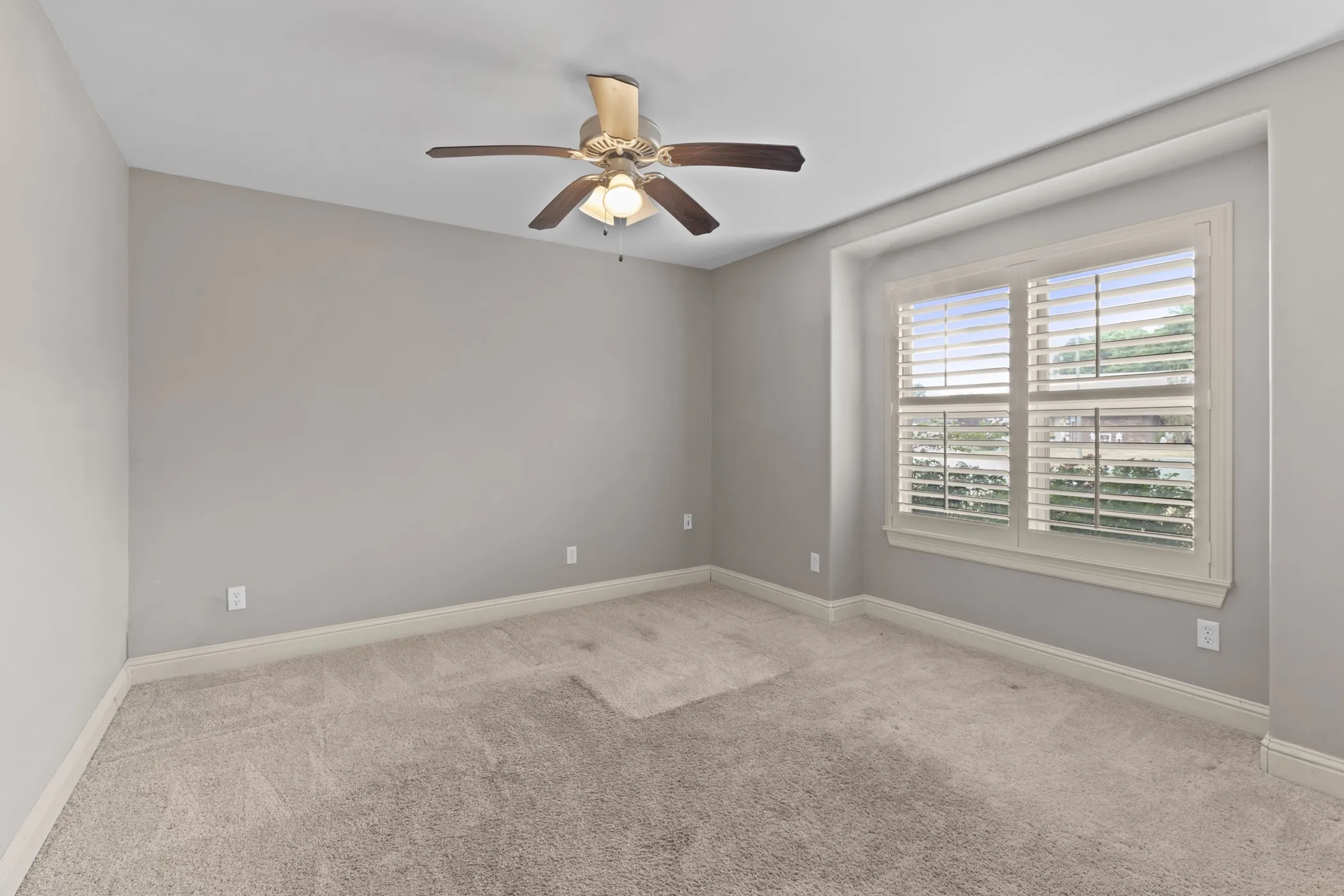 Neutral tone carpeting, light gray wall paint, window with white plantation shutters, white trim, and a ceiling fan with integrated lighting