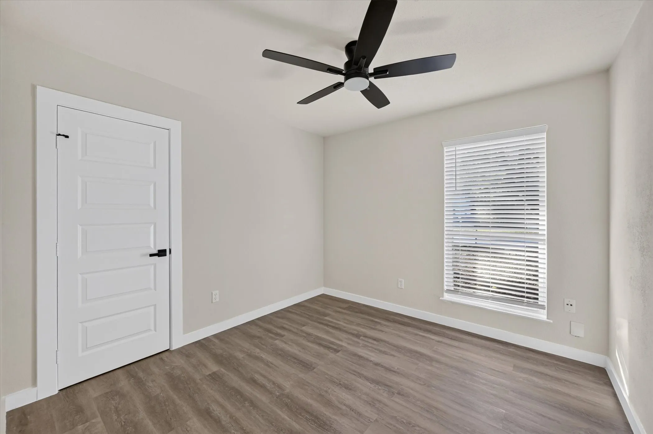 Wood-finish flooring, single window with blinds, ceiling fan, white paneled door with black hardware, and light-toned walls