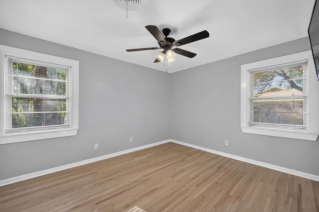 Spacious room featuring light gray walls, wood-finish flooring, two windows with blinds, and a ceiling fan with integrated lighting