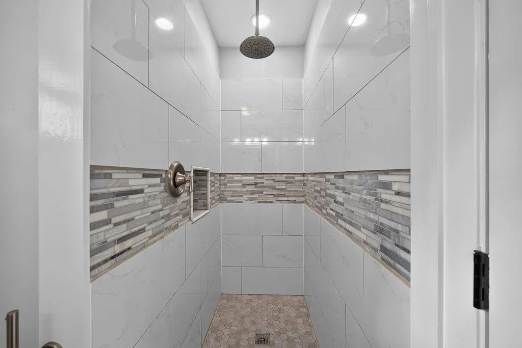 Spacious shower featuring full-height light-toned tile, decorative mosaic tile band, a built-in niche, and a rain shower head