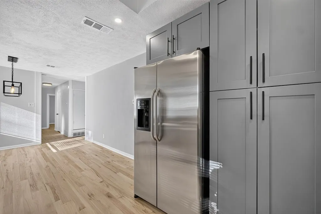 Full-height grey cabinetry with matte black hardware, stainless steel refrigerator, recessed lighting, wood-finish flooring, and a modern ceiling light fixture