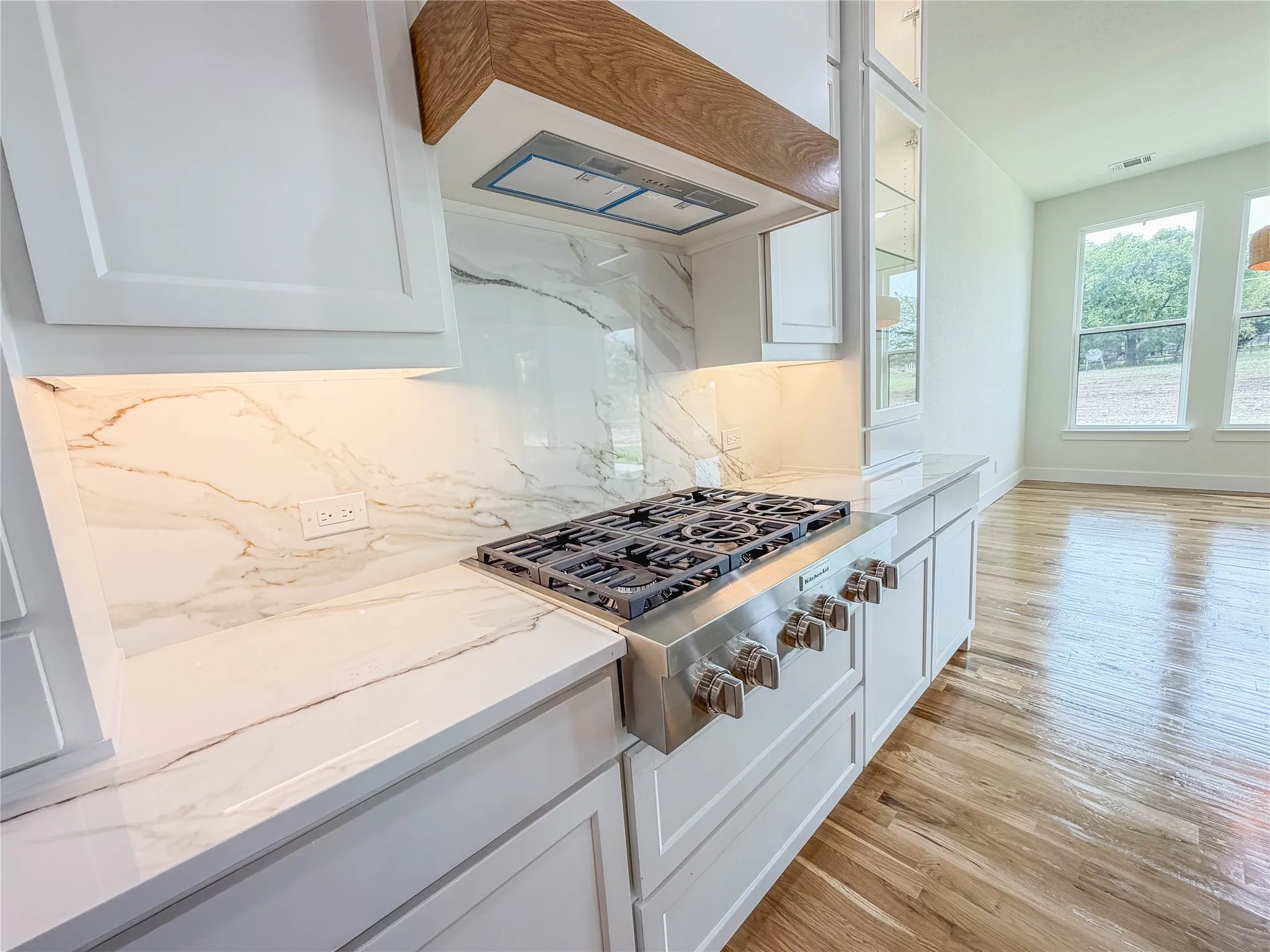 Gourmet kitchen featuring a built-in gas range, wood-finish range hood, white shaker cabinetry, and a marbled backsplash